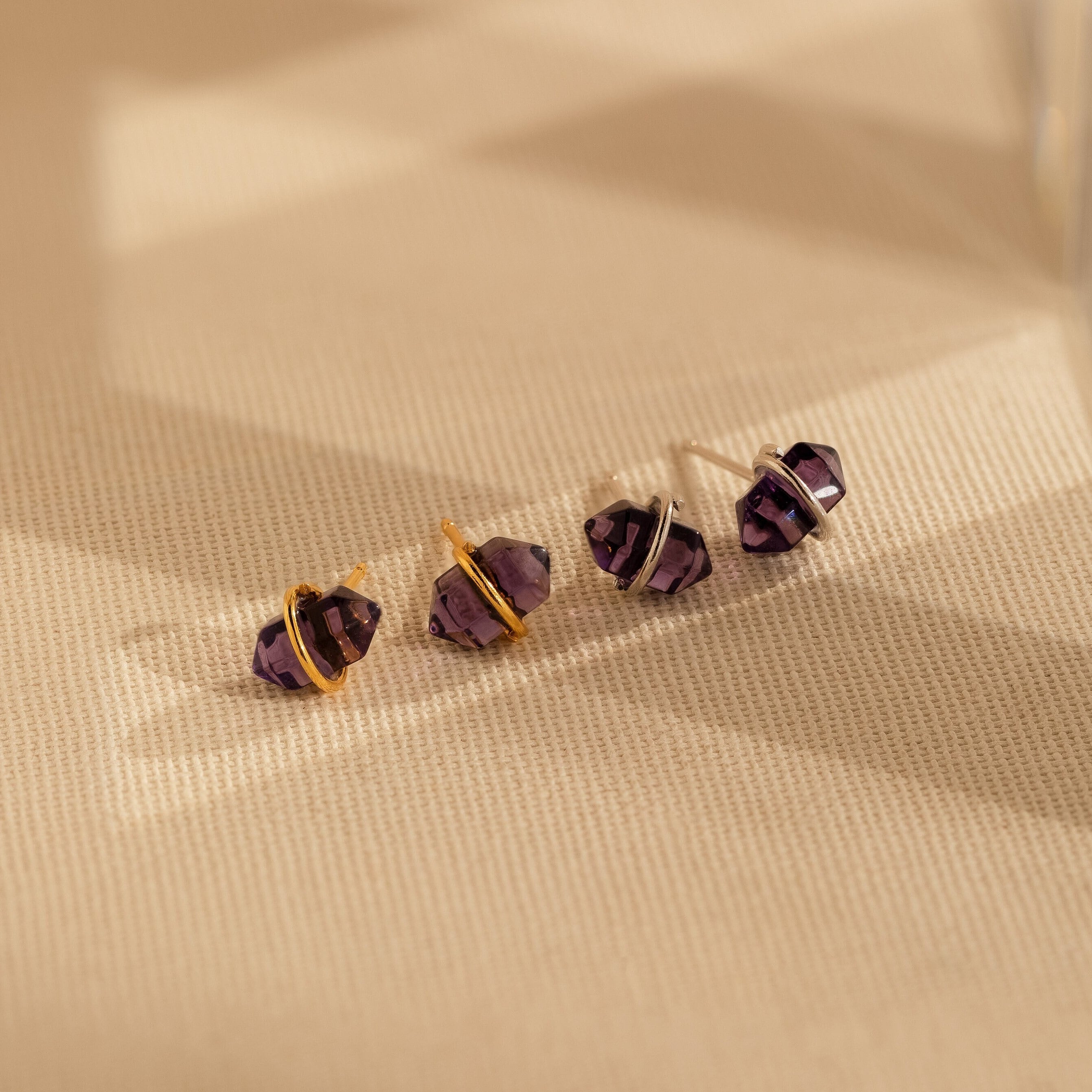 Four Amethyst Herkimer Studs, featuring gold and silver bands, are elegantly displayed on a textured beige surface.