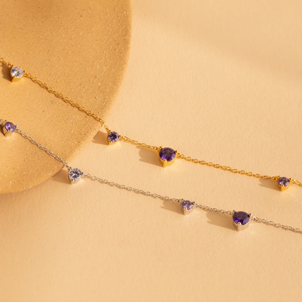The Amethyst Station Necklace features delicate chains adorned with purple amethyst and clear gemstones, set against a beige background—a stylish gift for any occasion.