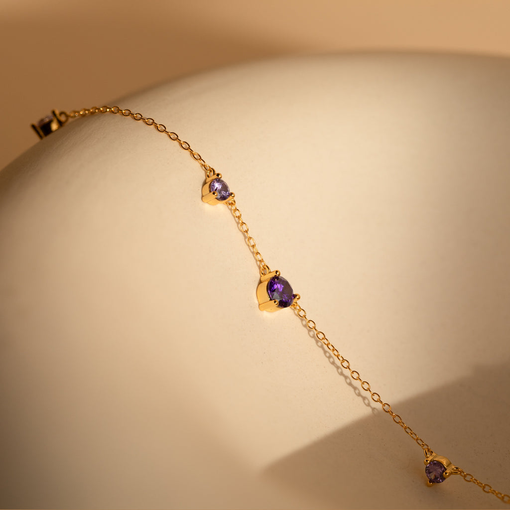 The Amethyst Station Necklace features a delicate gold chain with three amethyst charms, elegantly displayed on a smooth beige surface—a stylish gift for any occasion.