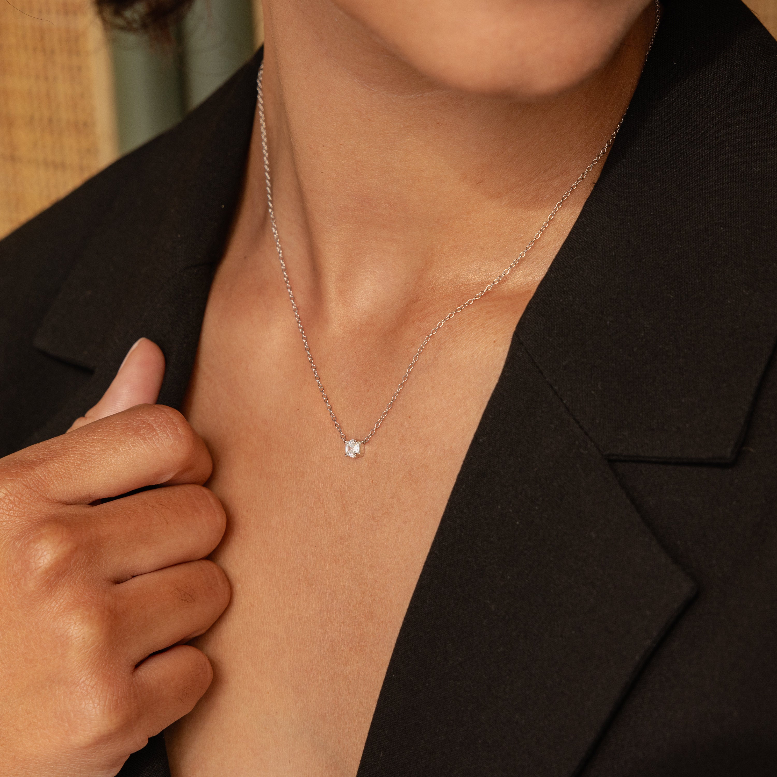 A person wearing a black blazer and the Hexagon Diamond Necklace, a delicate geometric piece featuring a small hexagon diamond pendant.