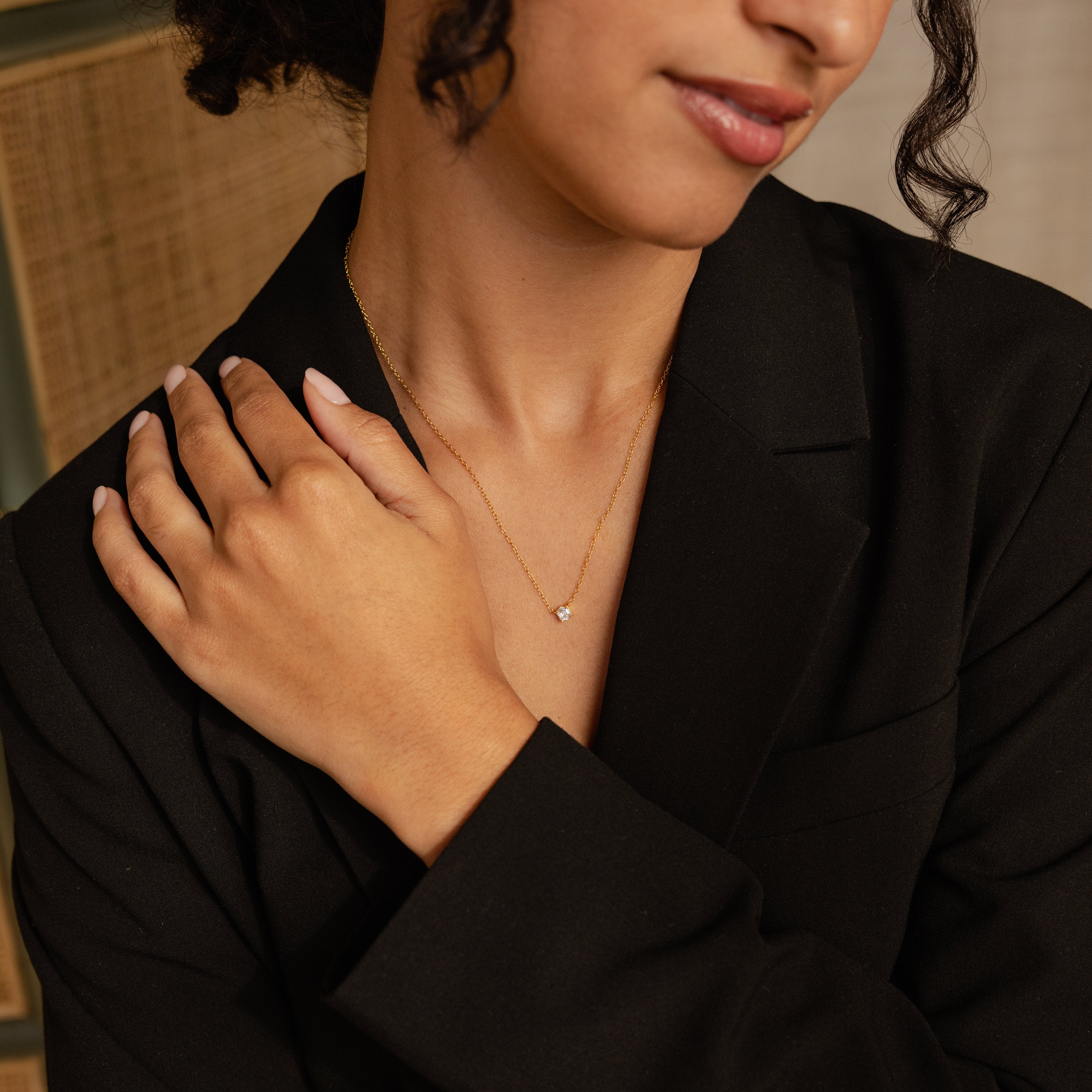 A woman in a black blazer touches her collarbone while wearing the Hexagon Diamond Necklace, a delicate gold piece featuring a small hexagonal pendant.
