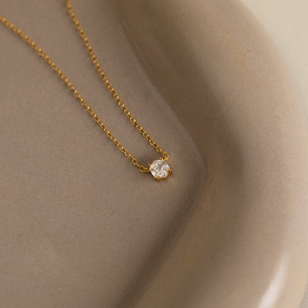 The Hexagon Diamond Necklace features a gold chain with a single rose-cut diamond pendant, showcased on a smooth beige ceramic dish.