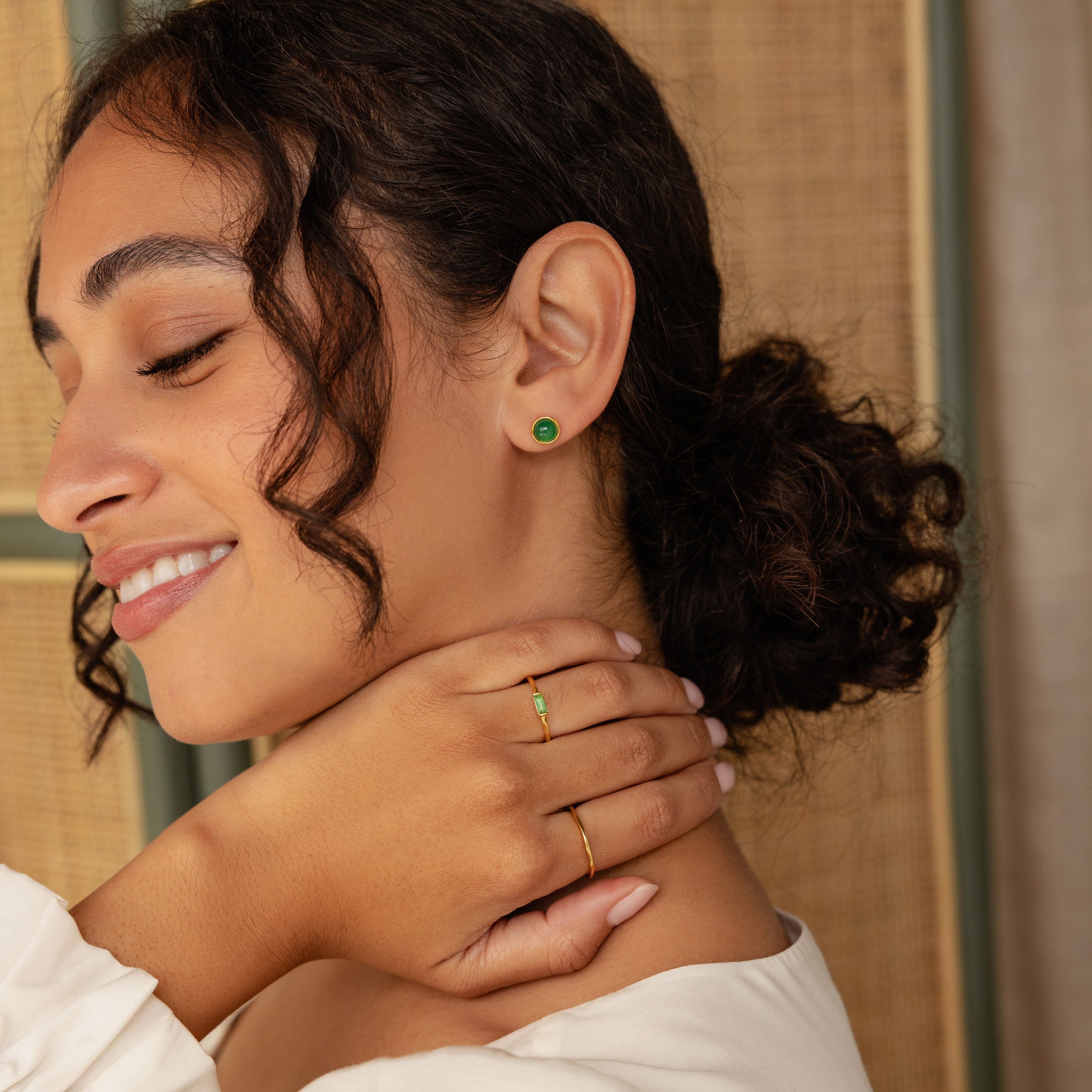 A woman with curly hair smiles, wearing Manifest Gemstone Studs and a gold ring with a green stone, her hand near her neck.