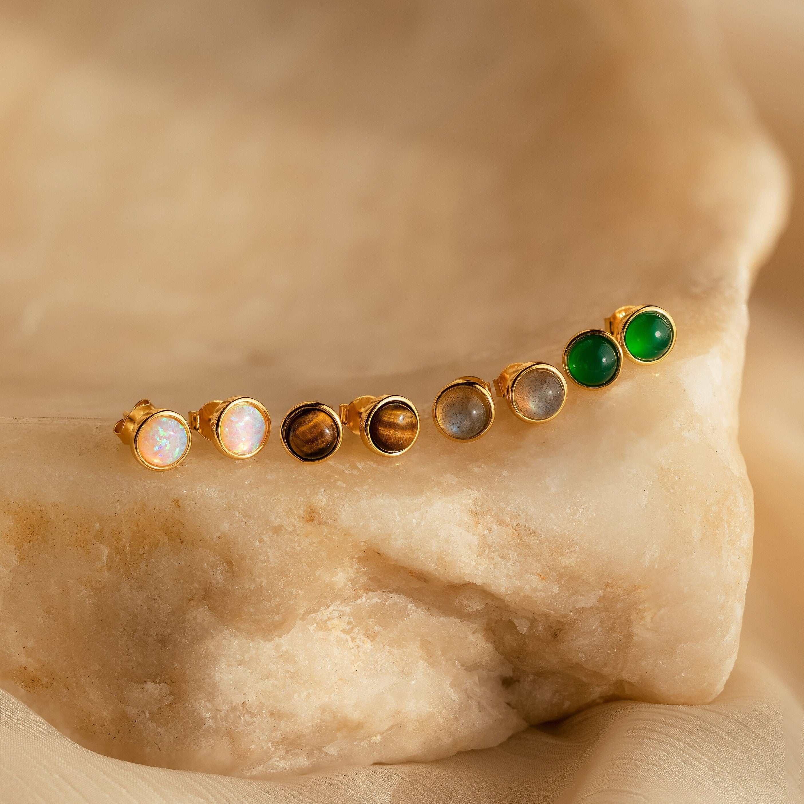Five pairs of Manifest Gemstone Studs, featuring gold settings and stones like labradorite and tigers eye, are displayed on a textured beige stone surface.
