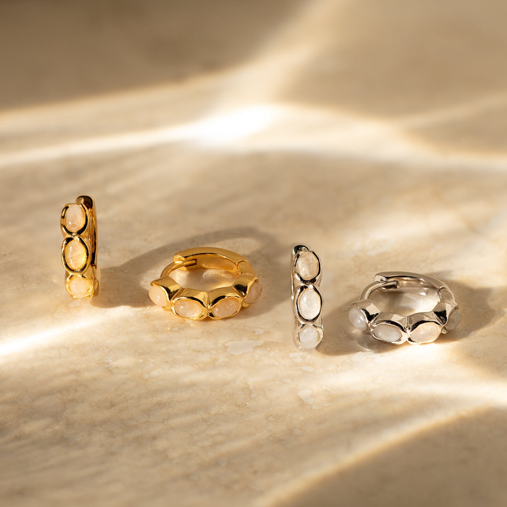 Two Moonstone Huggies earrings in gold and two in silver, each featuring oval details, are displayed on a marbled beige surface under sunlight.