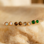 Four pairs of Manifest Gemstone Studs, featuring Labradorite and Tigers Eye earrings with colorful centers, are displayed on a beige stone surface.