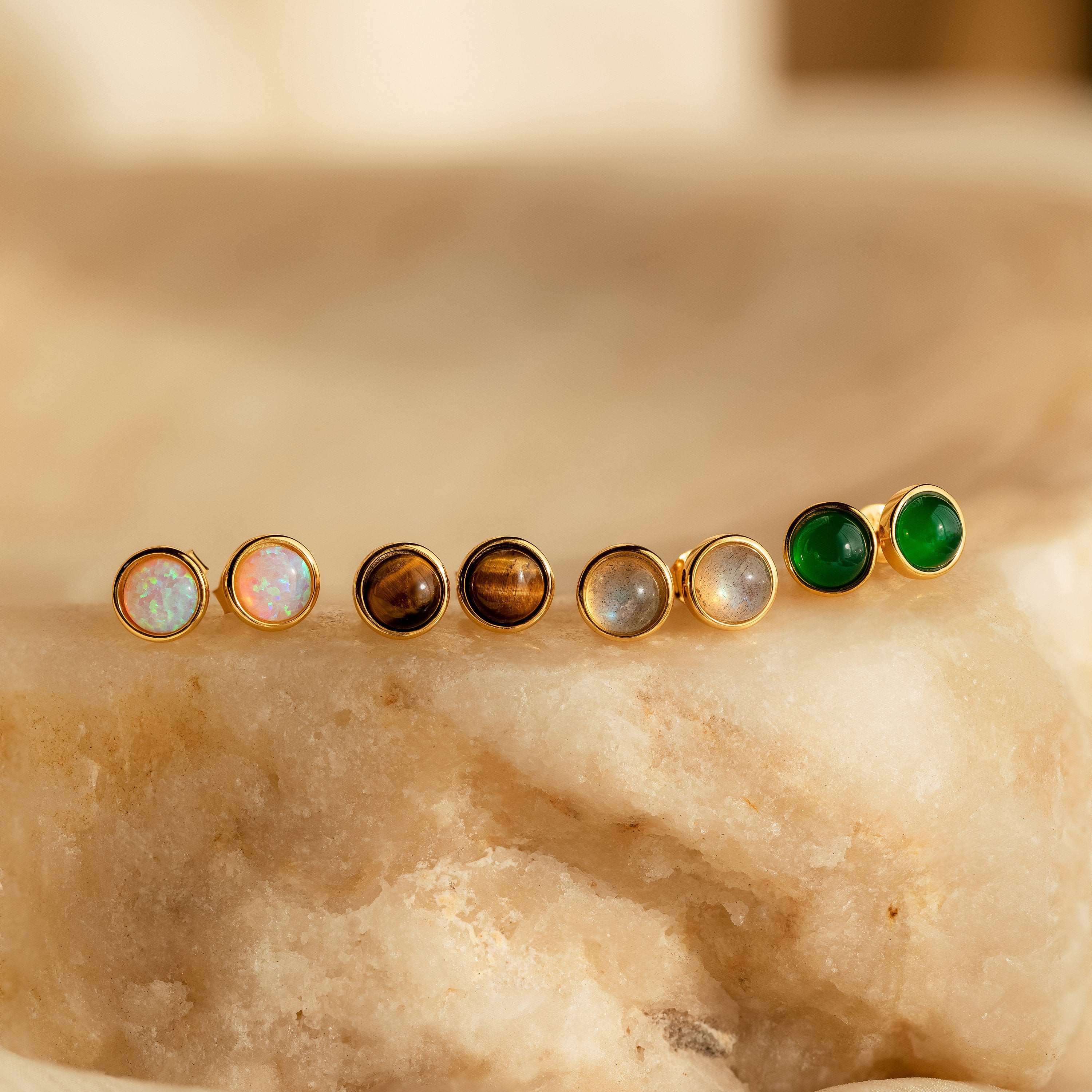 Four pairs of Manifest Gemstone Studs, featuring Labradorite and Tigers Eye earrings with colorful centers, are displayed on a beige stone surface.
