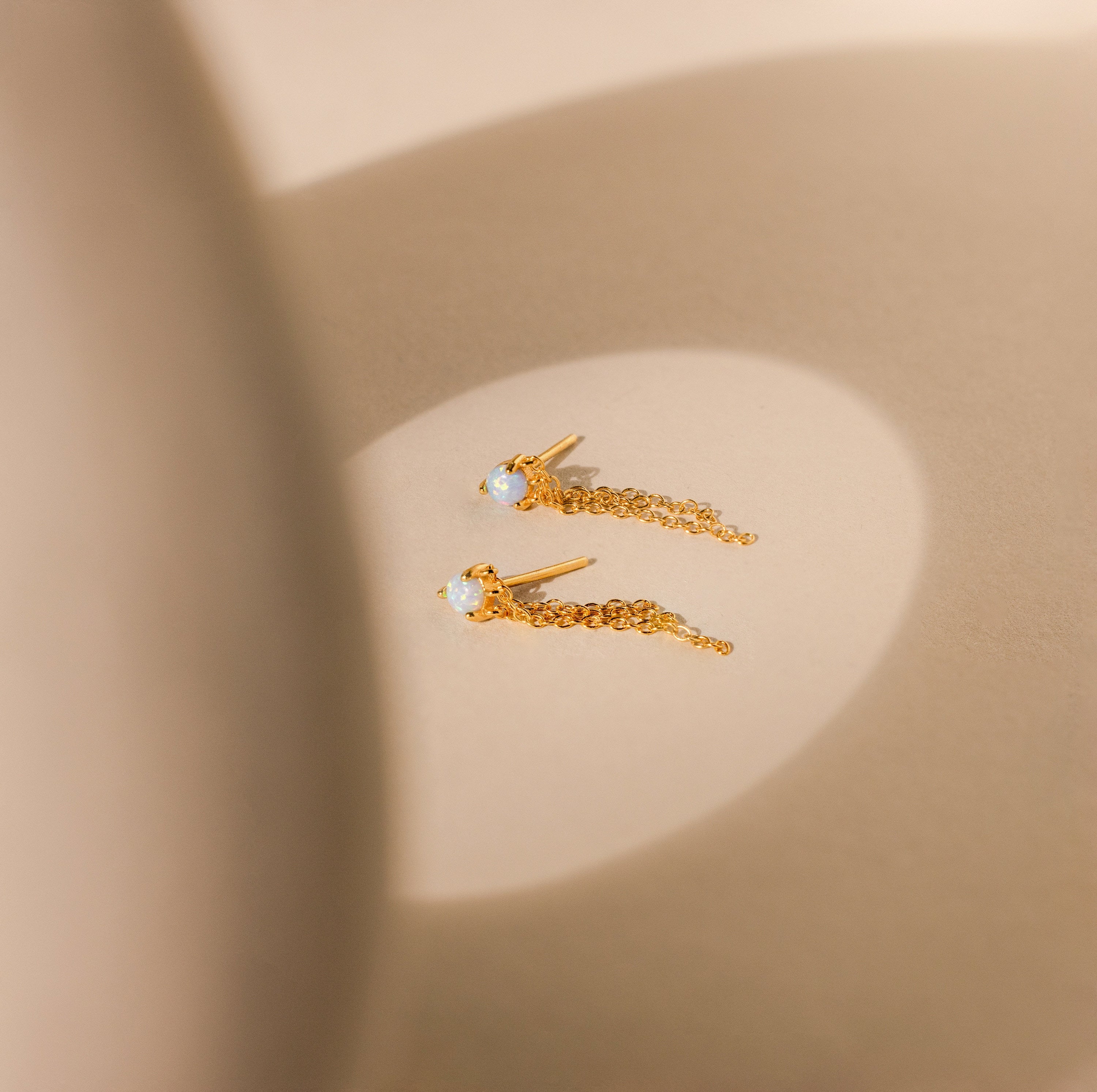Opal Chain Earrings are displayed on a beige surface in soft, natural light, beautifully highlighting their delicate design and elegance.