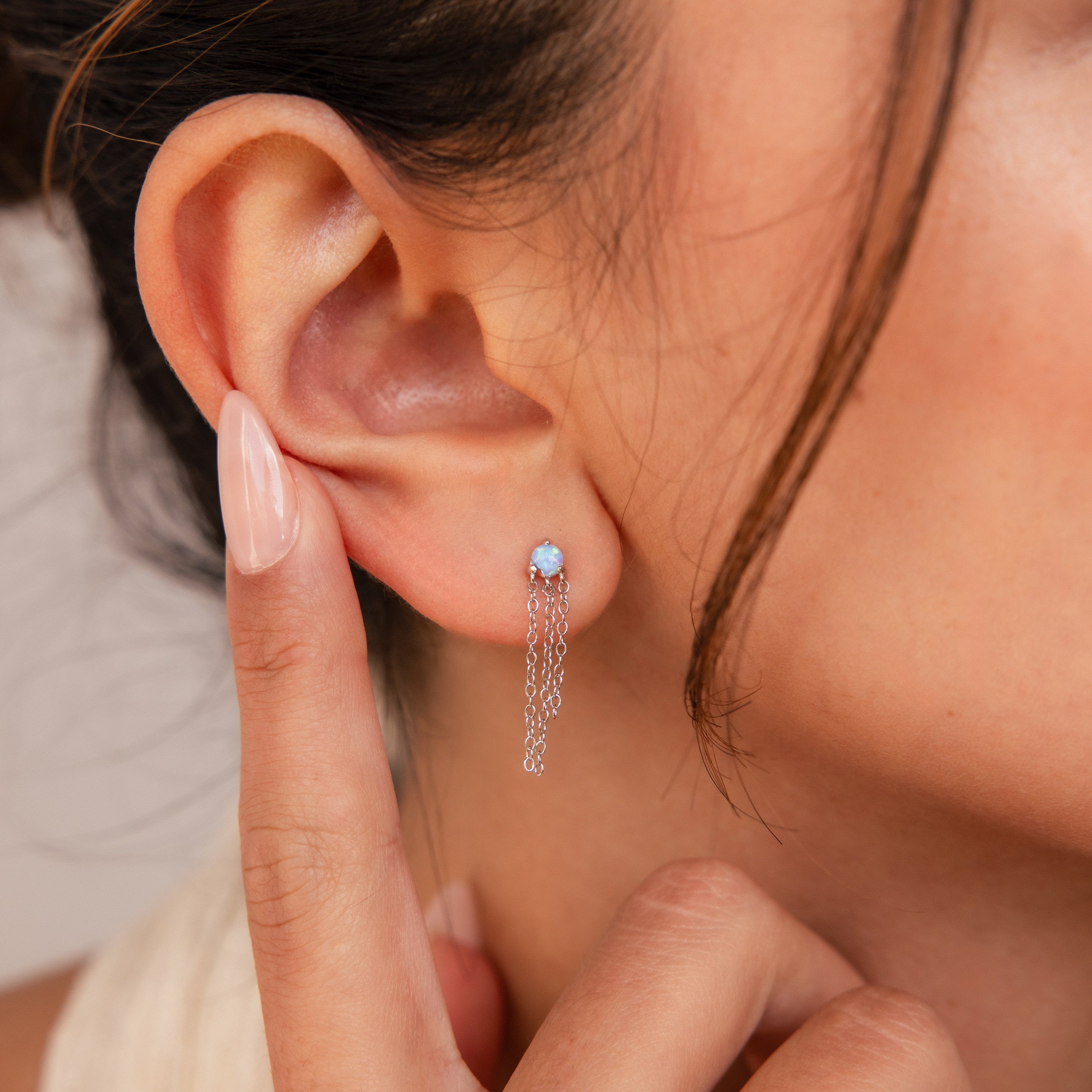 A woman touches her ear while wearing Opal Chain Earrings, featuring blue gemstones and dangling silver chains—a striking addition to any jewelry collection.