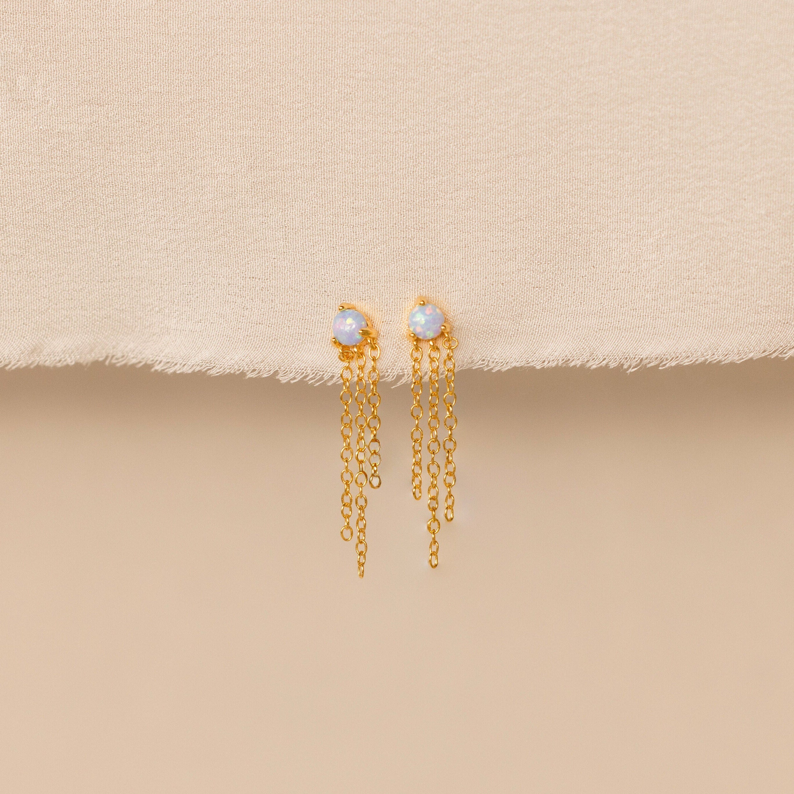 Opal Chain Earrings with opal studs and delicate dangling chains, displayed on a beige fabric background—an eye-catching piece from our newest jewelry collection.