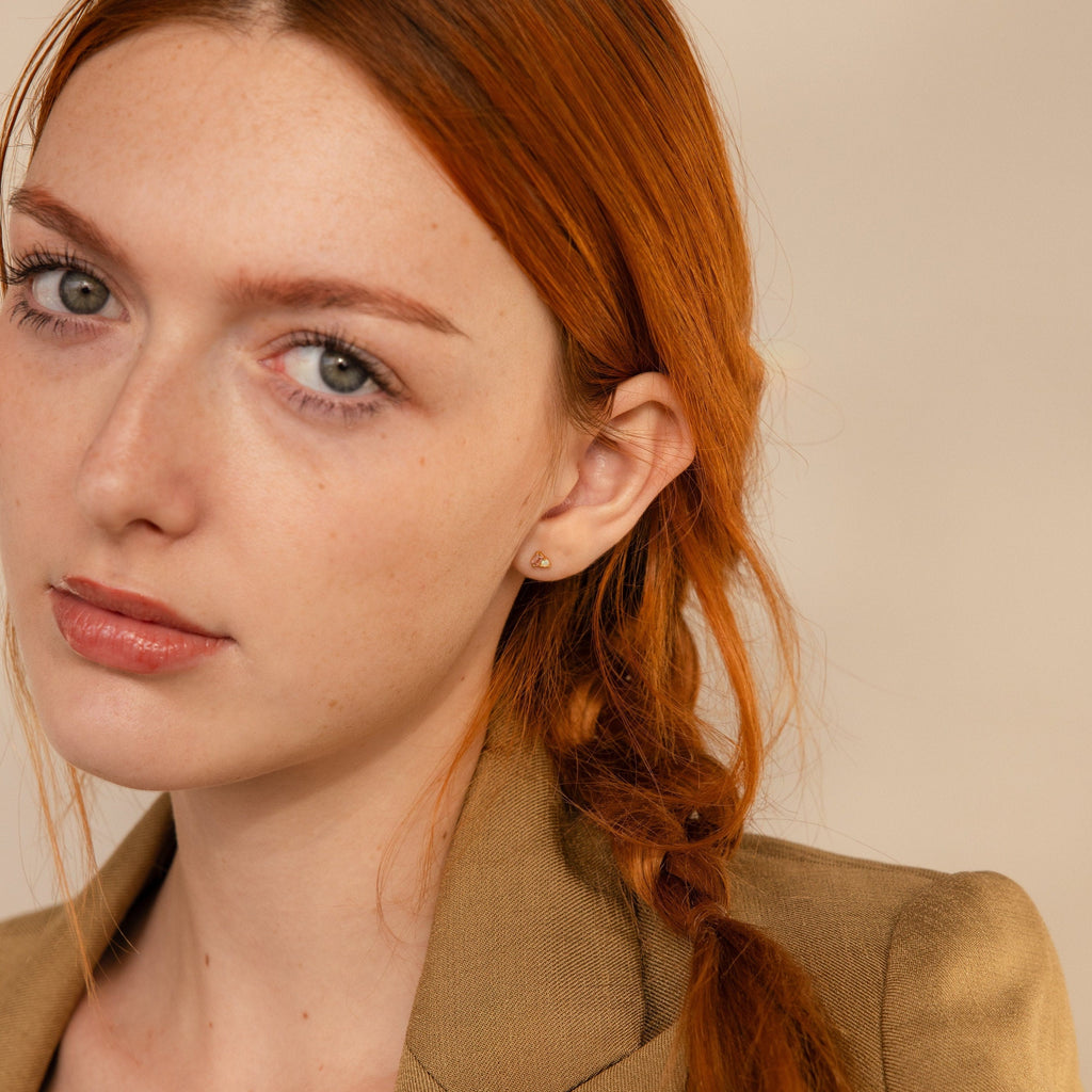 A woman with long red hair in a braid, wearing a tan blazer and elegant Pink Baguette Opal Studs, looks at the camera with a neutral expression.