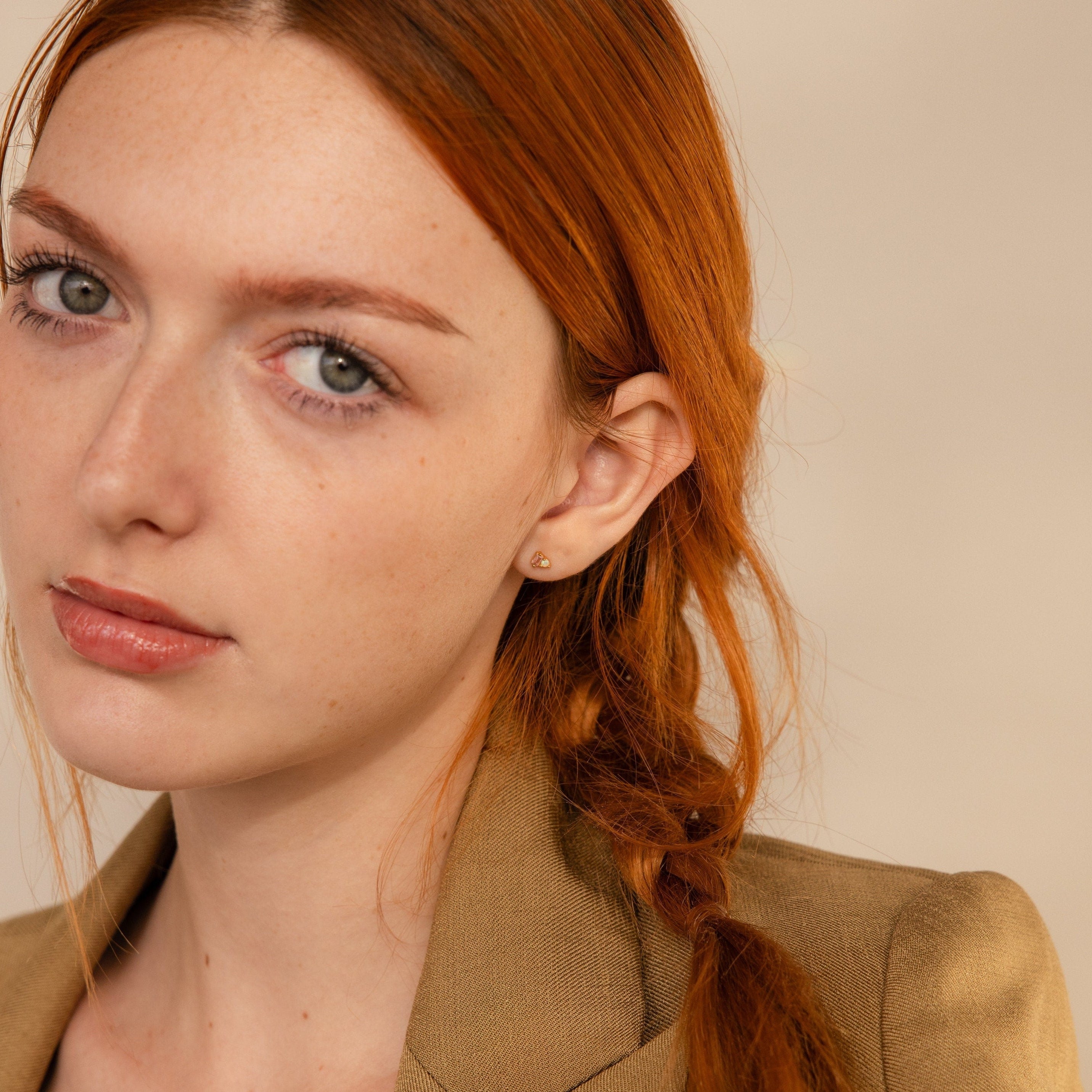 A woman with long red hair in a braid, wearing a tan blazer and elegant Pink Baguette Opal Studs, looks at the camera with a neutral expression.