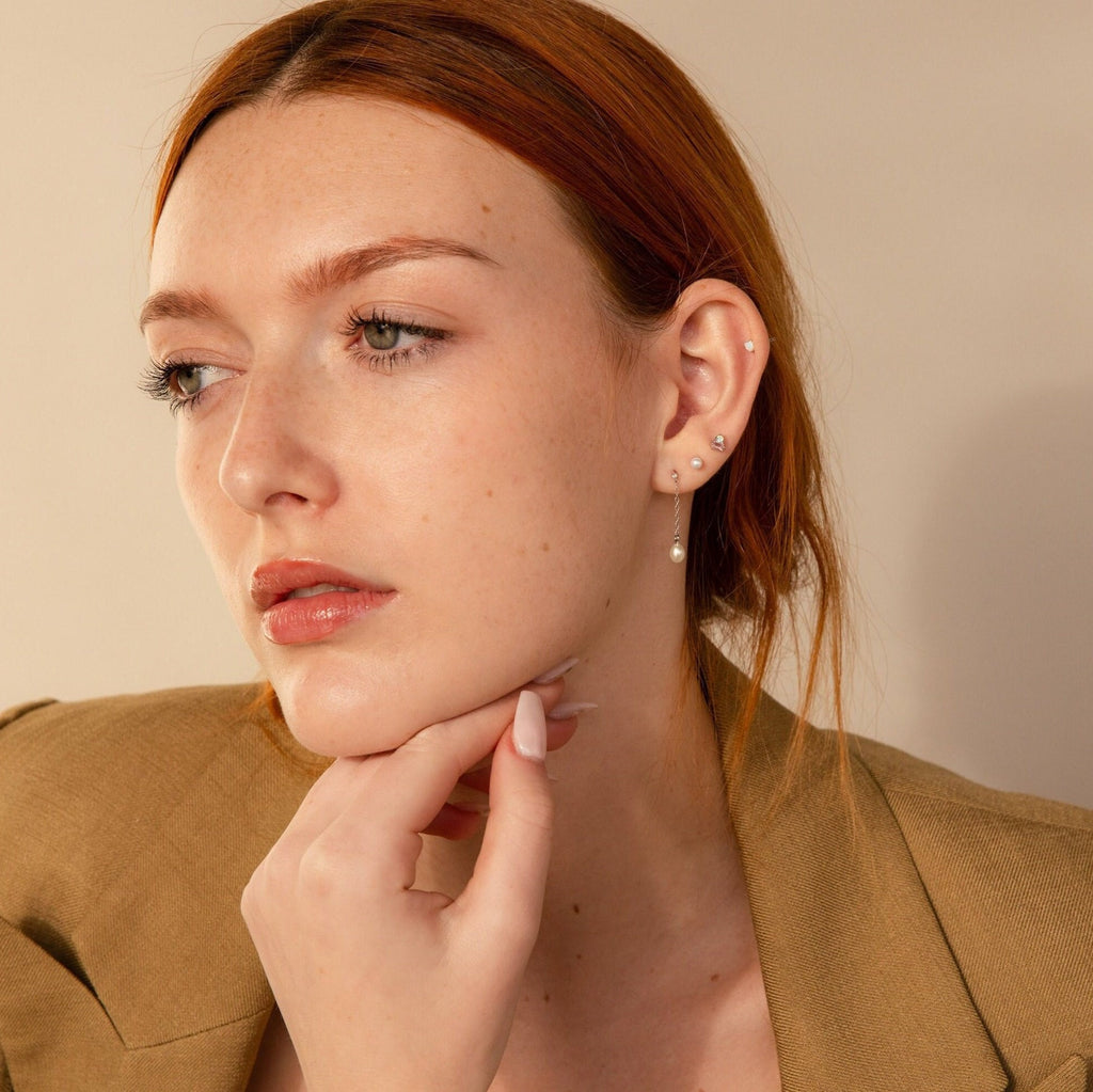 A woman with red hair and multiple ear piercings, dressed in a brown blazer and stylish Pink Baguette Opal Studs, rests her chin thoughtfully on her hand.