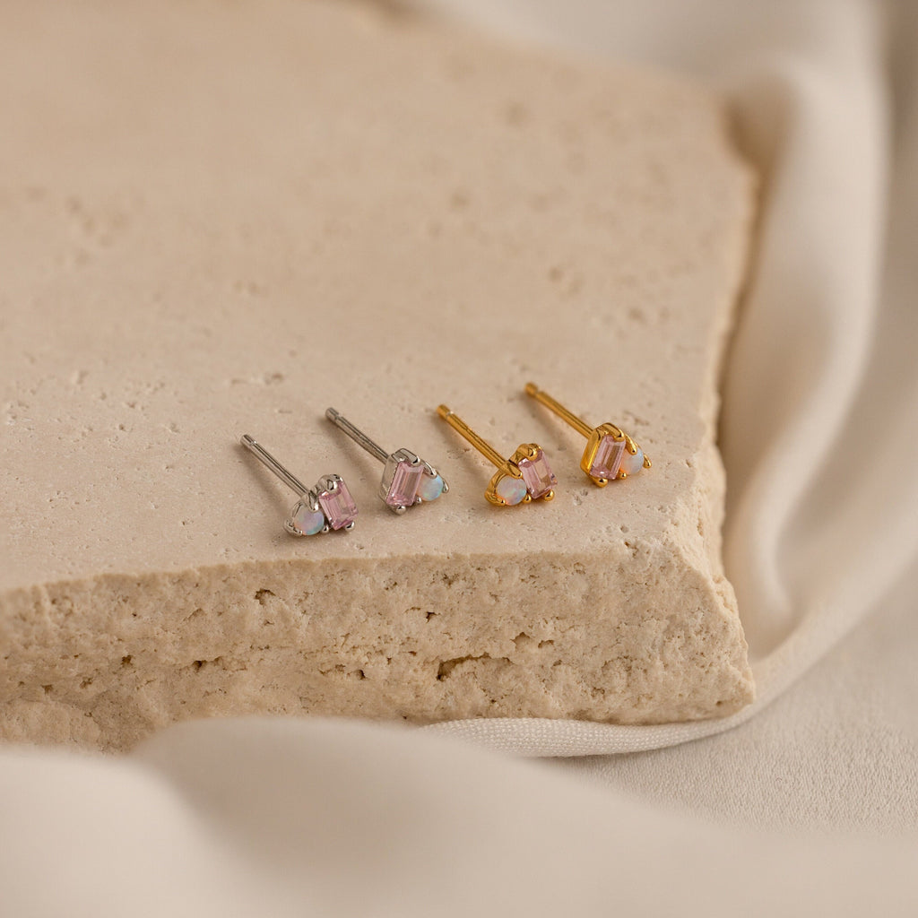 Two pairs of stud earrings—one silver Pink Baguette Opal Studs and one gold with pink gemstones—rest on a stone slab. Perfect as bridal gifts or for adding a touch of elegance to any look.