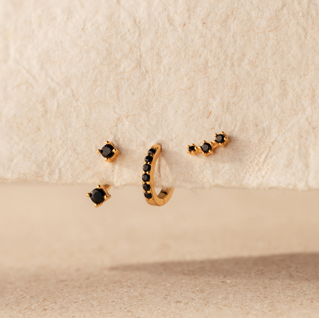The Black Diamond Earrings Set, featuring a climber stud and gemstone huggie hoop, is displayed on a textured beige surface.