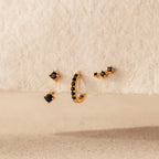 The Black Diamond Earrings Set, featuring a climber stud and gemstone huggie hoop, is displayed on a textured beige surface.