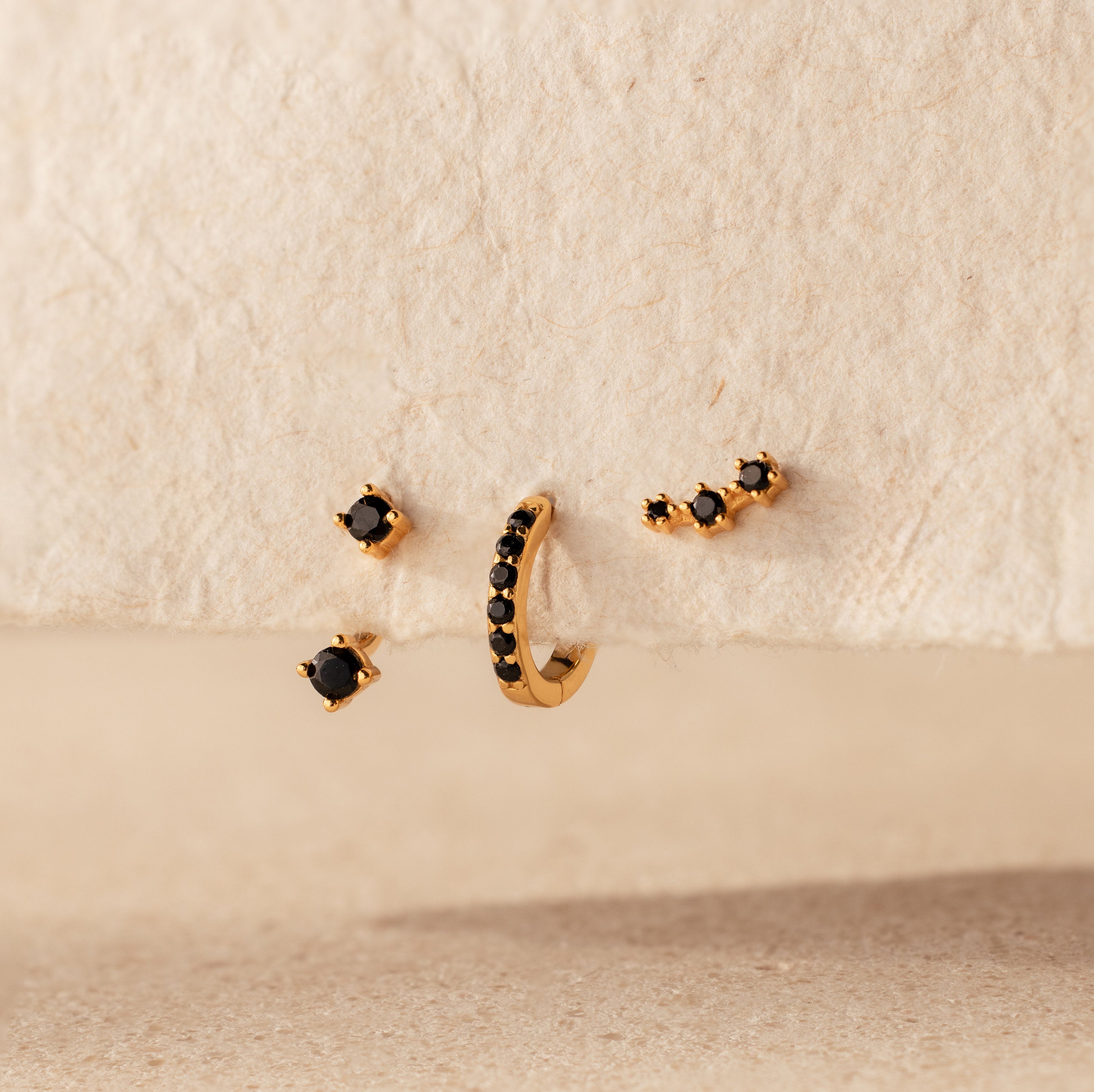 The Black Diamond Earrings Set, featuring a climber stud and gemstone huggie hoop, is displayed on a textured beige surface.