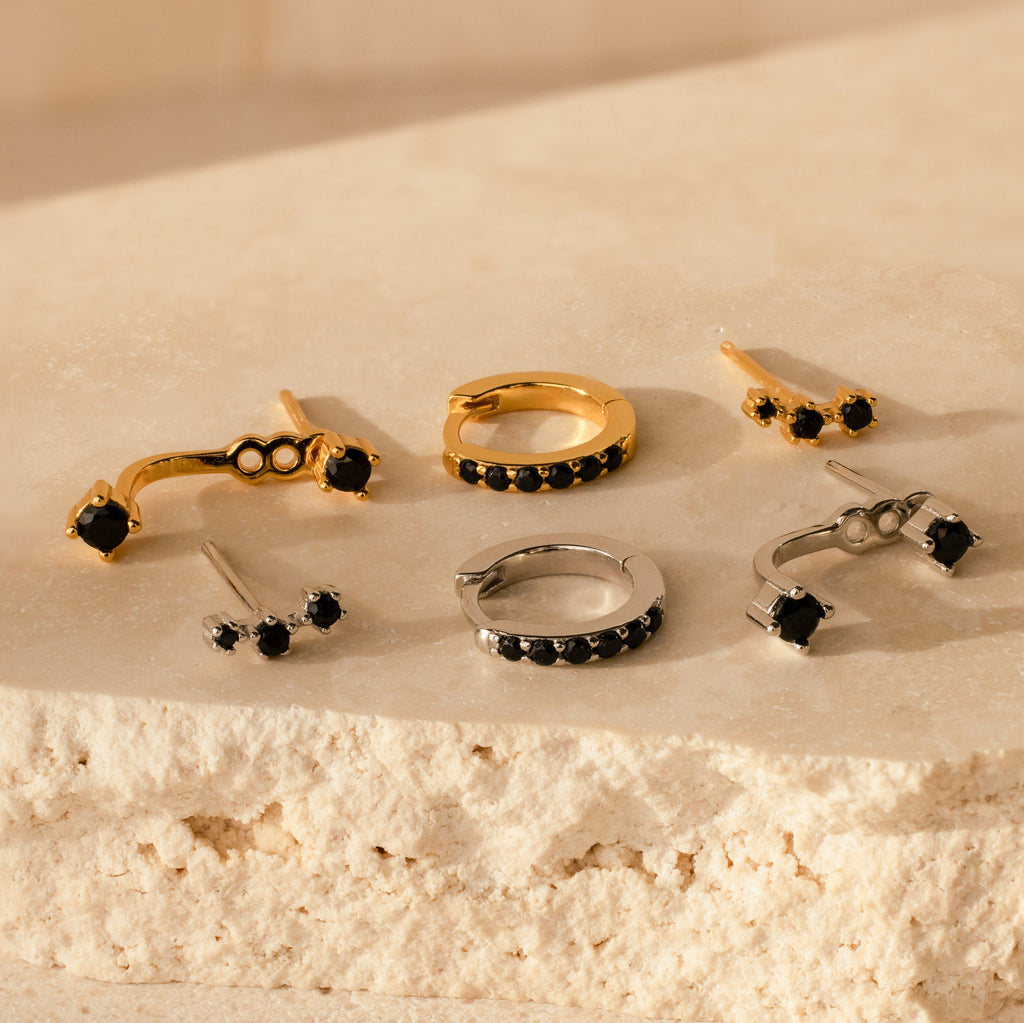 The Black Diamond Earrings Set features gold and silver earrings with black stones, displayed on a textured beige surface.