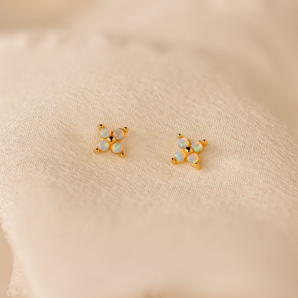 The Dainty Opal Stud Earrings Set features four small opal stones on gold settings, displayed against a soft cream fabric—ideal for those who love minimalist jewelry.