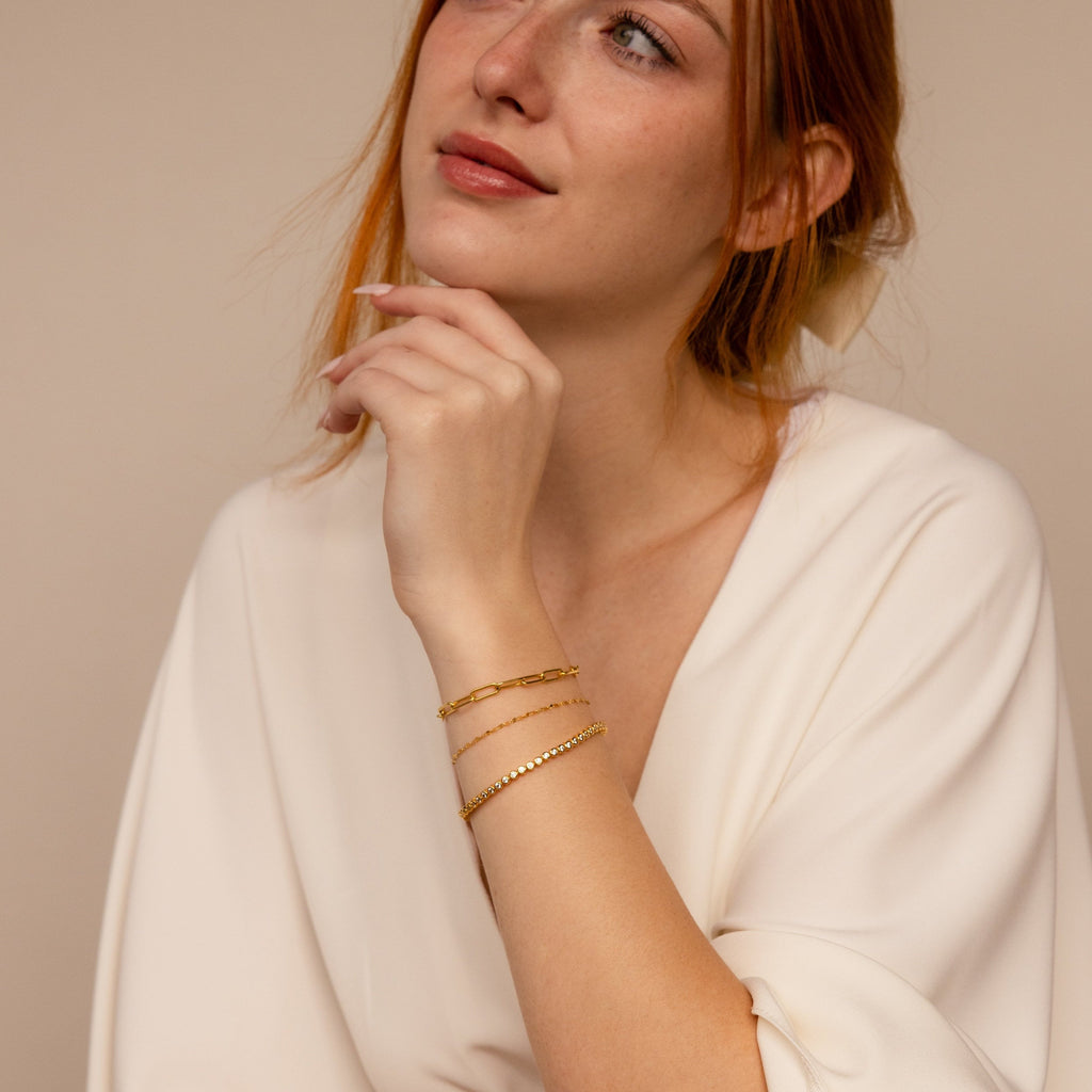 Woman in a white blouse wearing gold bracelets, including the Diamond Bezel Tennis Bracelet, gazes thoughtfully upward.