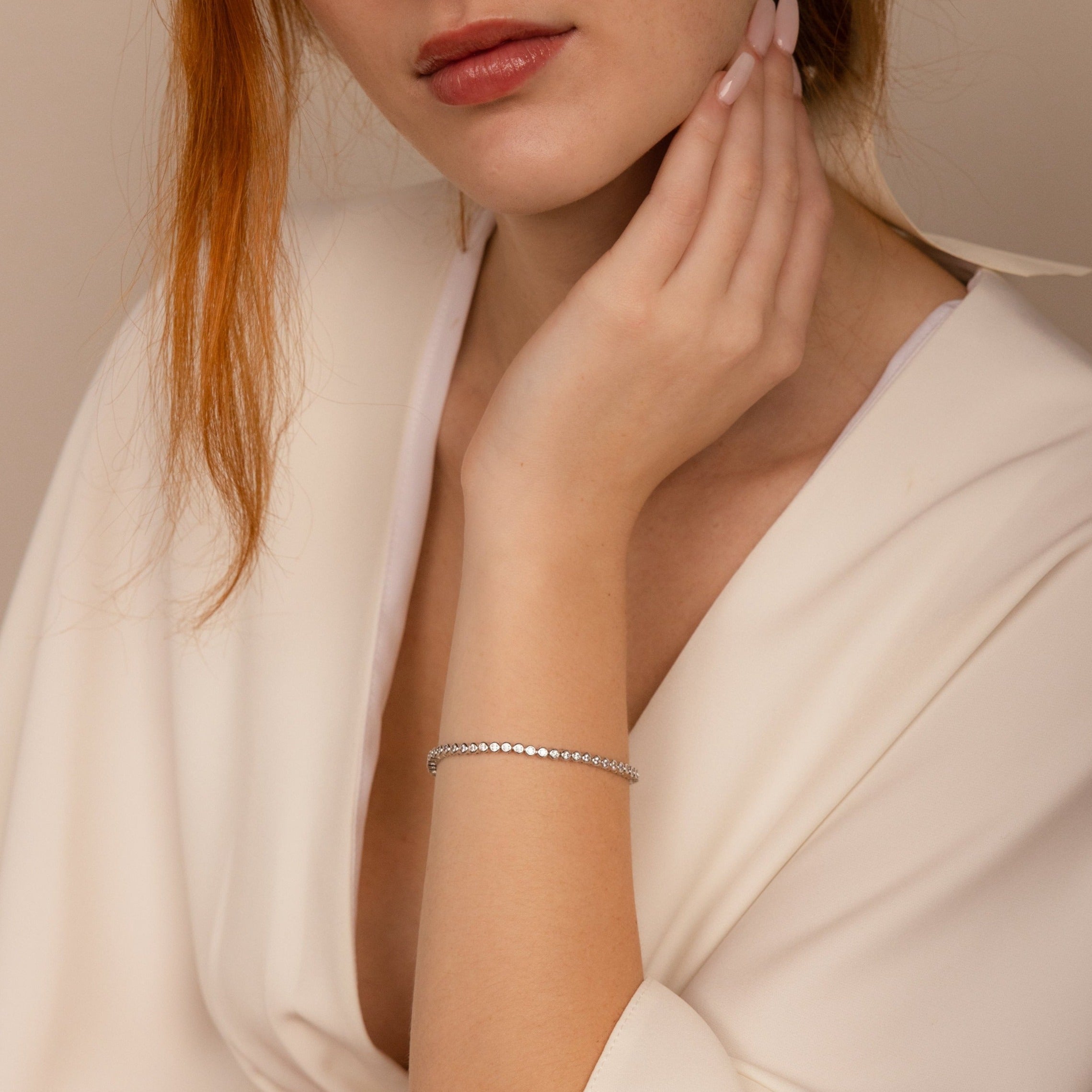 A woman in a cream blouse rests her hand on her face, elegantly showcasing the Diamond Bezel Tennis Bracelet.