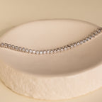 The Diamond Bezel Tennis Bracelet, featuring round diamonds, sits on a cream-colored textured dish—a minimalist gemstone bracelet and ideal personalized bridal gift.