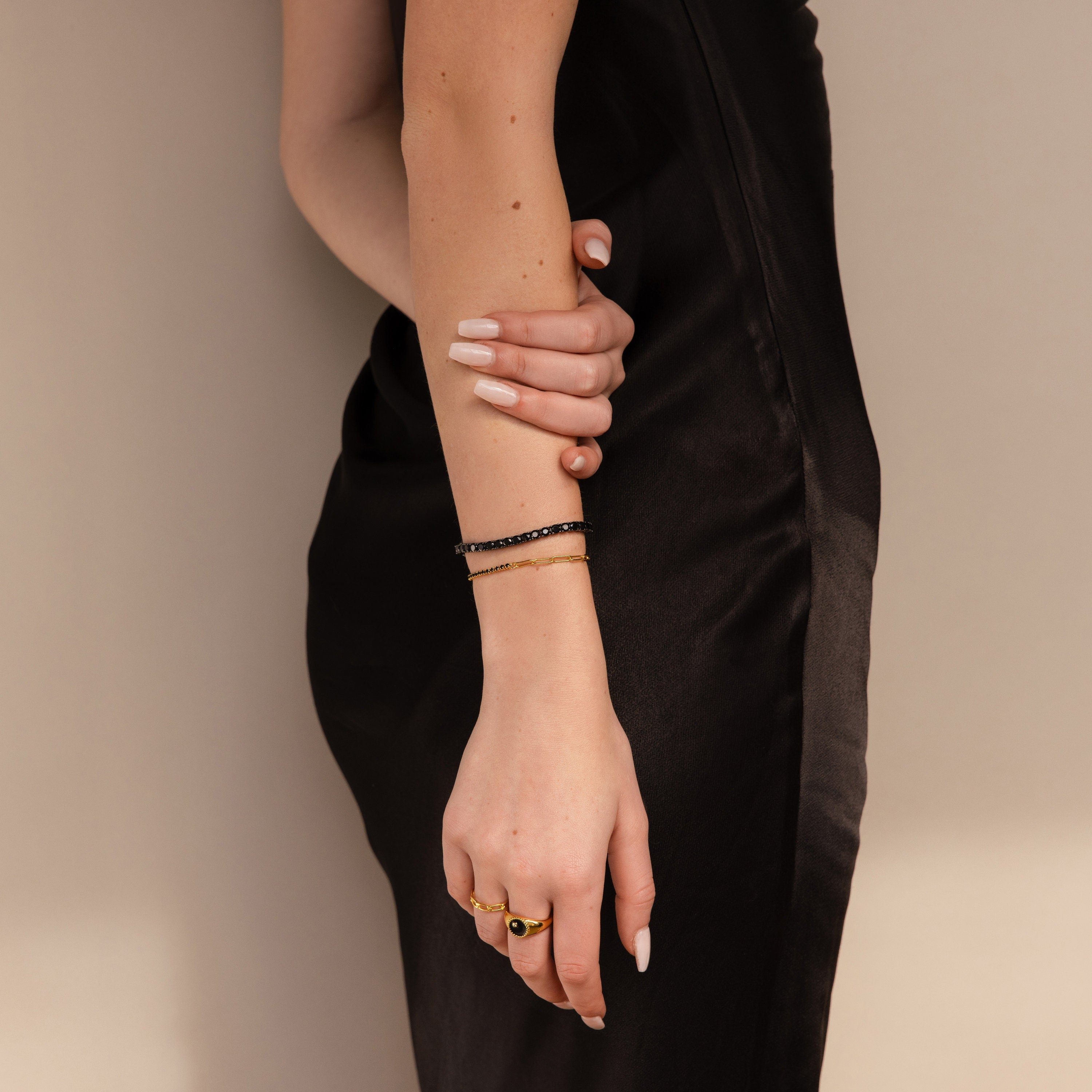A woman in a black dress, showcasing manicured nails, gold rings, and the elegant Onyx Tennis Bracelet as she gently holds her arm.