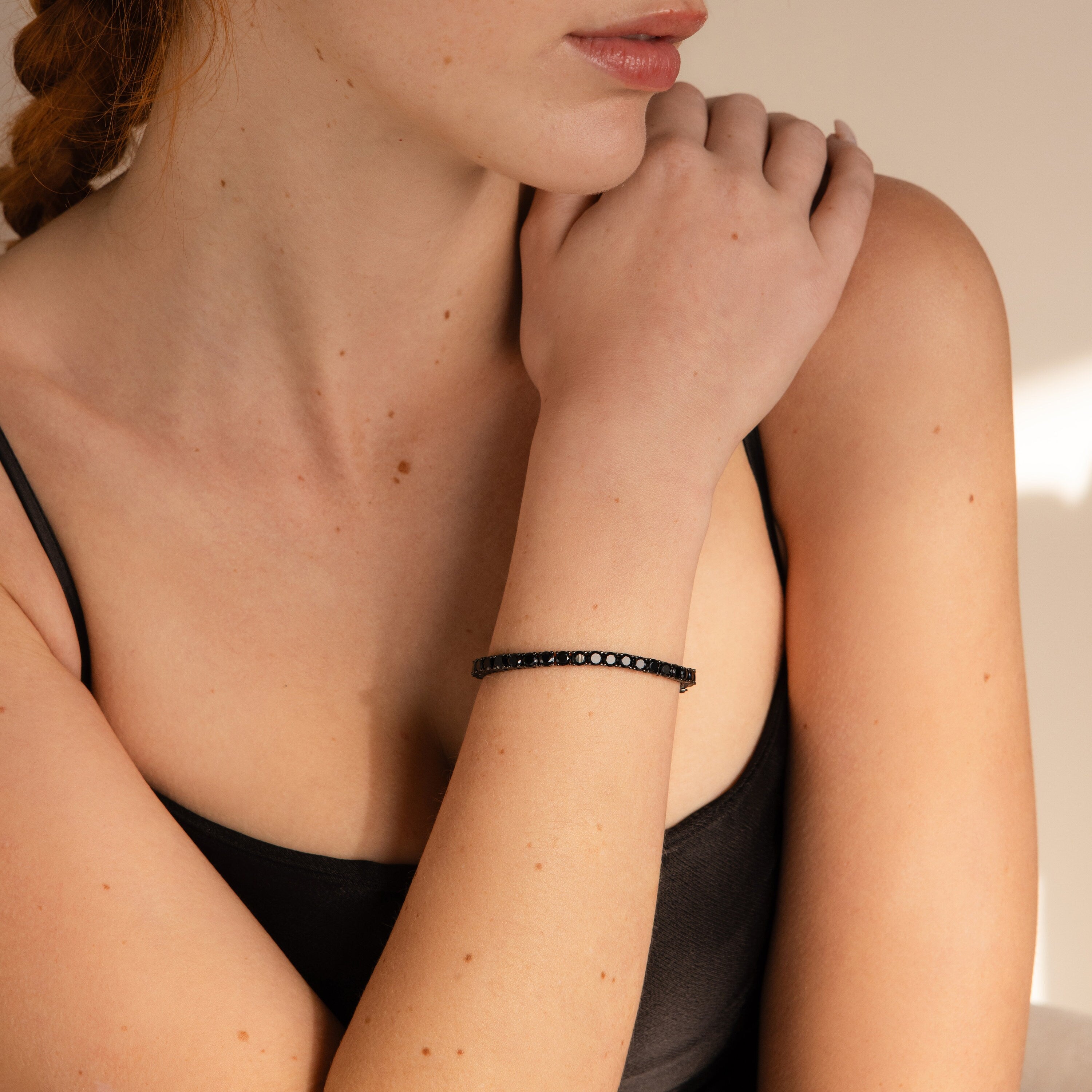 A woman in a black tank top, softly lit, rests her chin on her hand while wearing the stunning Onyx Tennis Bracelet.