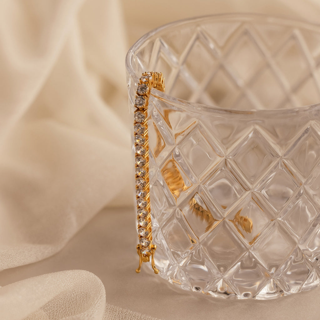 The Bold Diamond Tennis Bracelet is draped over a faceted glass cup on soft beige fabric—a striking, elegant accessory perfect for bridesmaid jewelry.