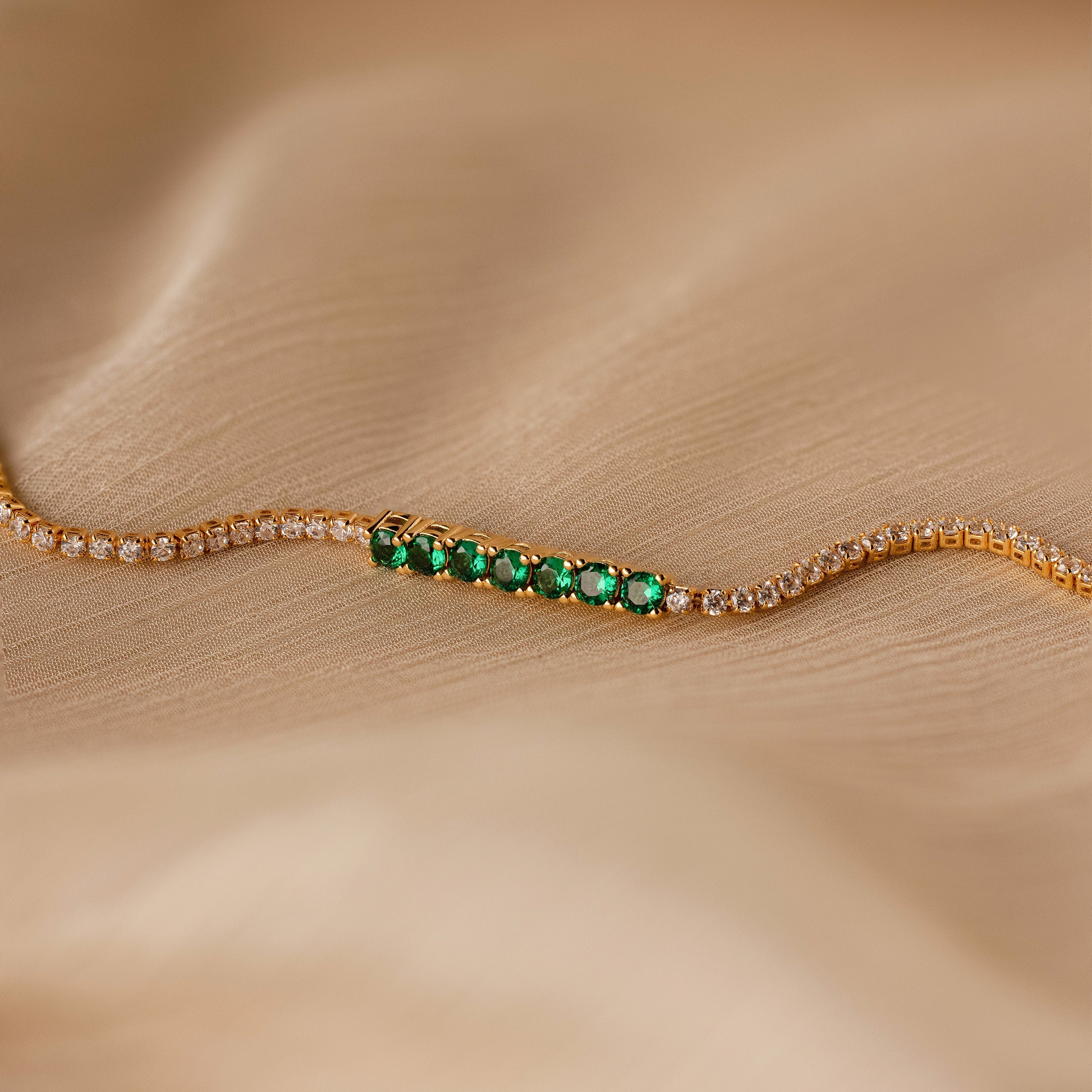The Emerald Diamond Tennis Bracelet, featuring sparkling diamonds and a row of emerald gemstones, is elegantly displayed on soft beige fabric.