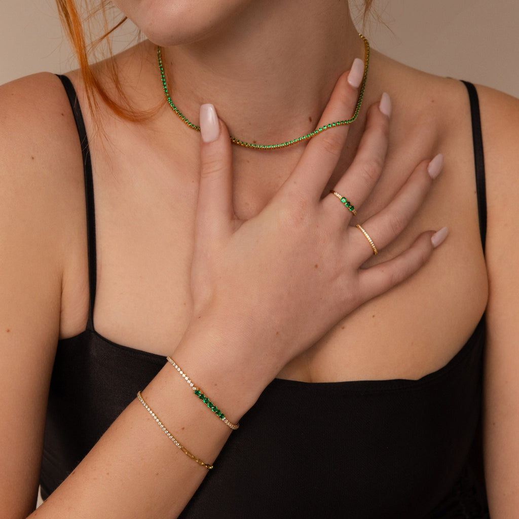 Woman in a black top, wearing gold and green jewelry—Emerald Diamond Tennis Bracelet, matching necklace and rings with diamond stones—and manicured nails.