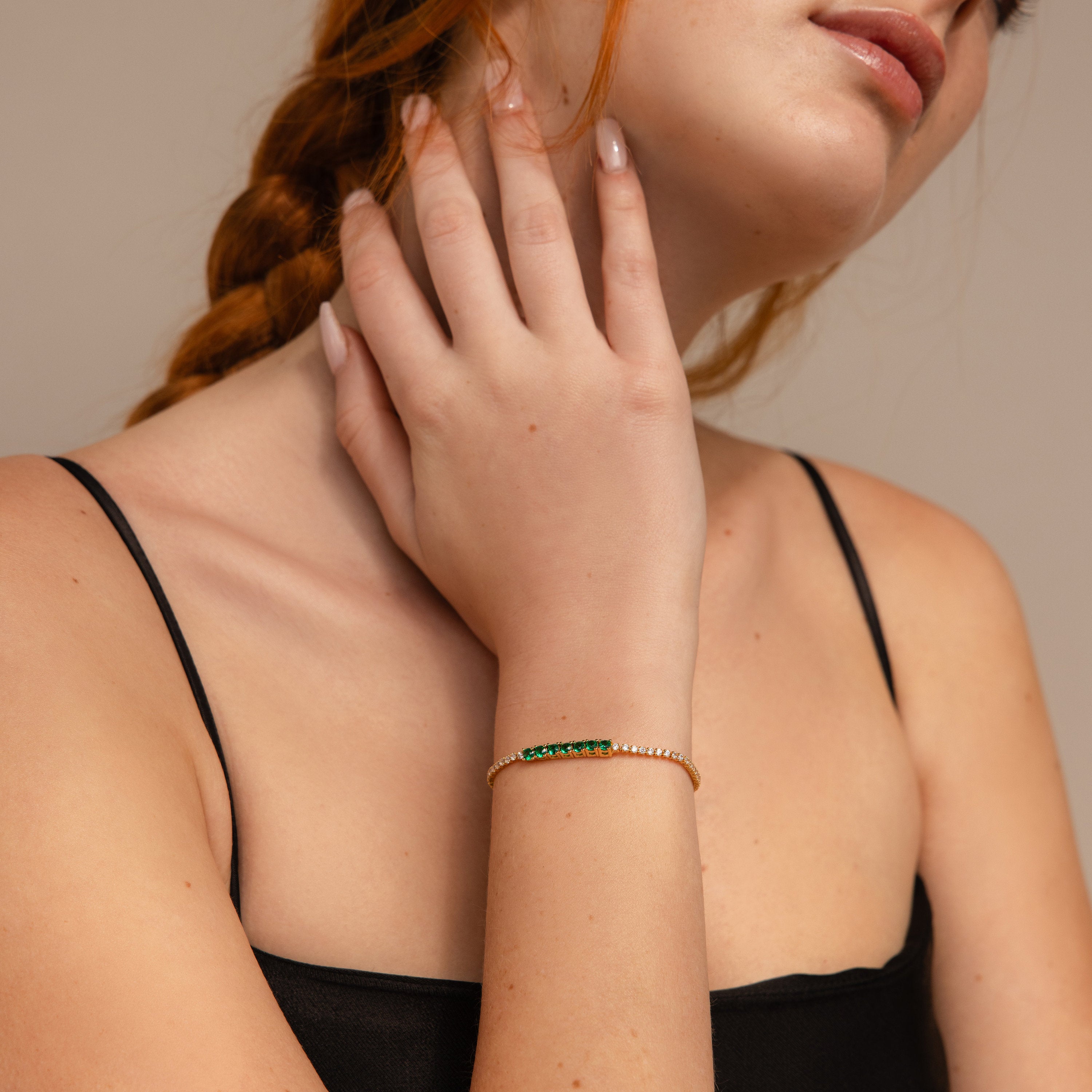 A woman with braided hair wearing a black top and an elegant Emerald Diamond Tennis Bracelet touches her neck with her left hand.