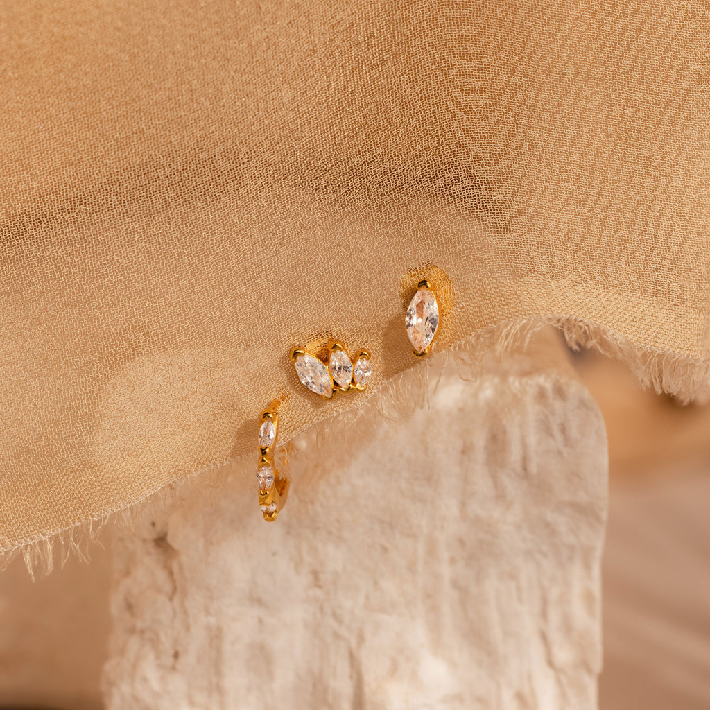The Marquise Diamond Earrings Set, ideal as bridesmaid gifts, features gold hoops with clear gemstones and is displayed on a textured stone and beige fabric background.