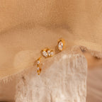 The Marquise Diamond Earrings Set, ideal as bridesmaid gifts, features gold hoops with clear gemstones and is displayed on a textured stone and beige fabric background.