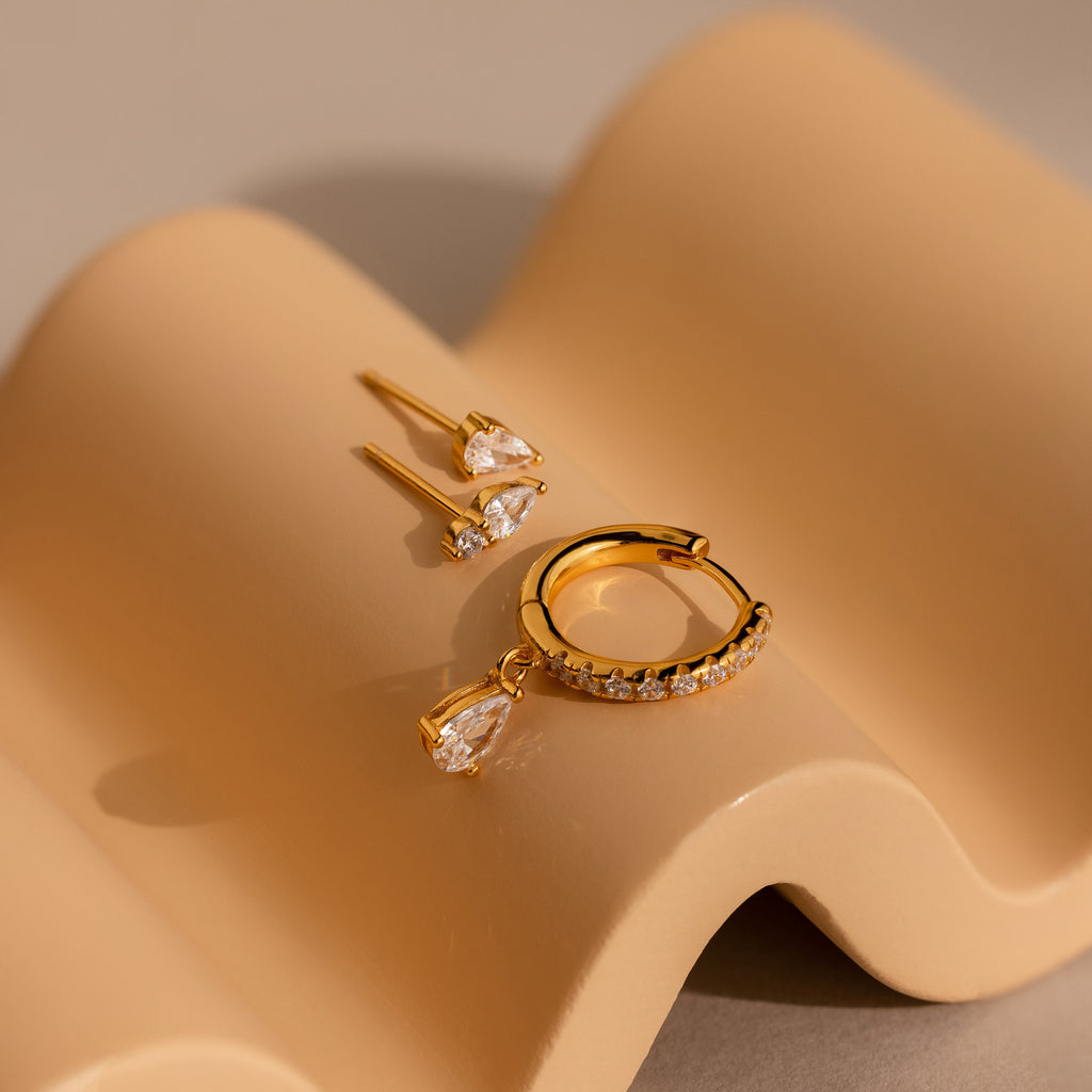 The Diamond Drop Earrings Set, featuring teardrop studs and a crystal pave hoop earring, is displayed on a wavy beige surface.