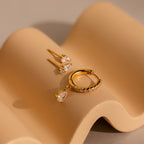 The Diamond Drop Earrings Set, featuring teardrop studs and a crystal pave hoop earring, is displayed on a wavy beige surface.