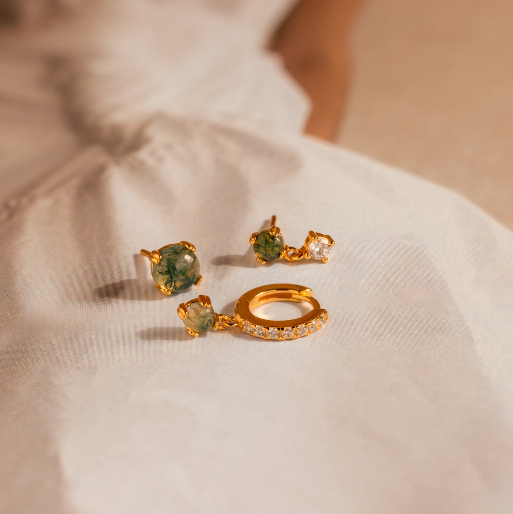 The Agate Earrings Set features two gold earrings with green stones and a single gold hoop with clear stones, arranged on a light fabric surface for an elegant look.
