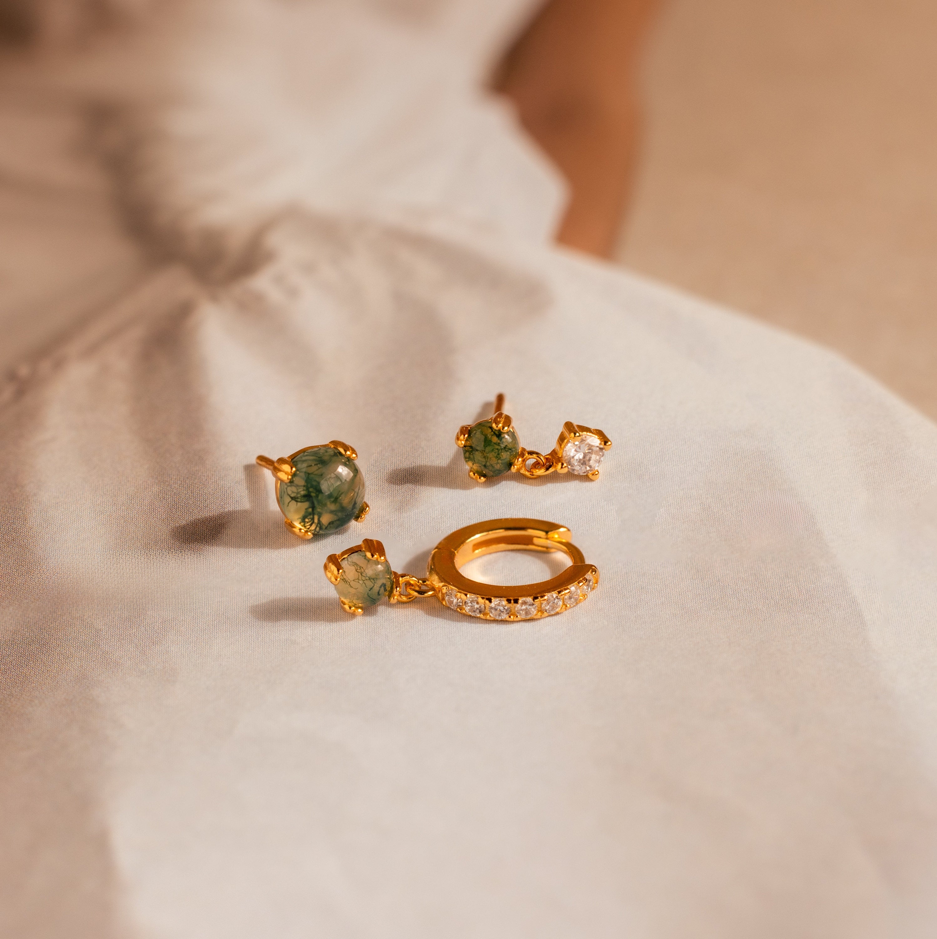 The Agate Earrings Set features two gold earrings with green stones and a single gold hoop with clear stones, arranged on a light fabric surface for an elegant look.