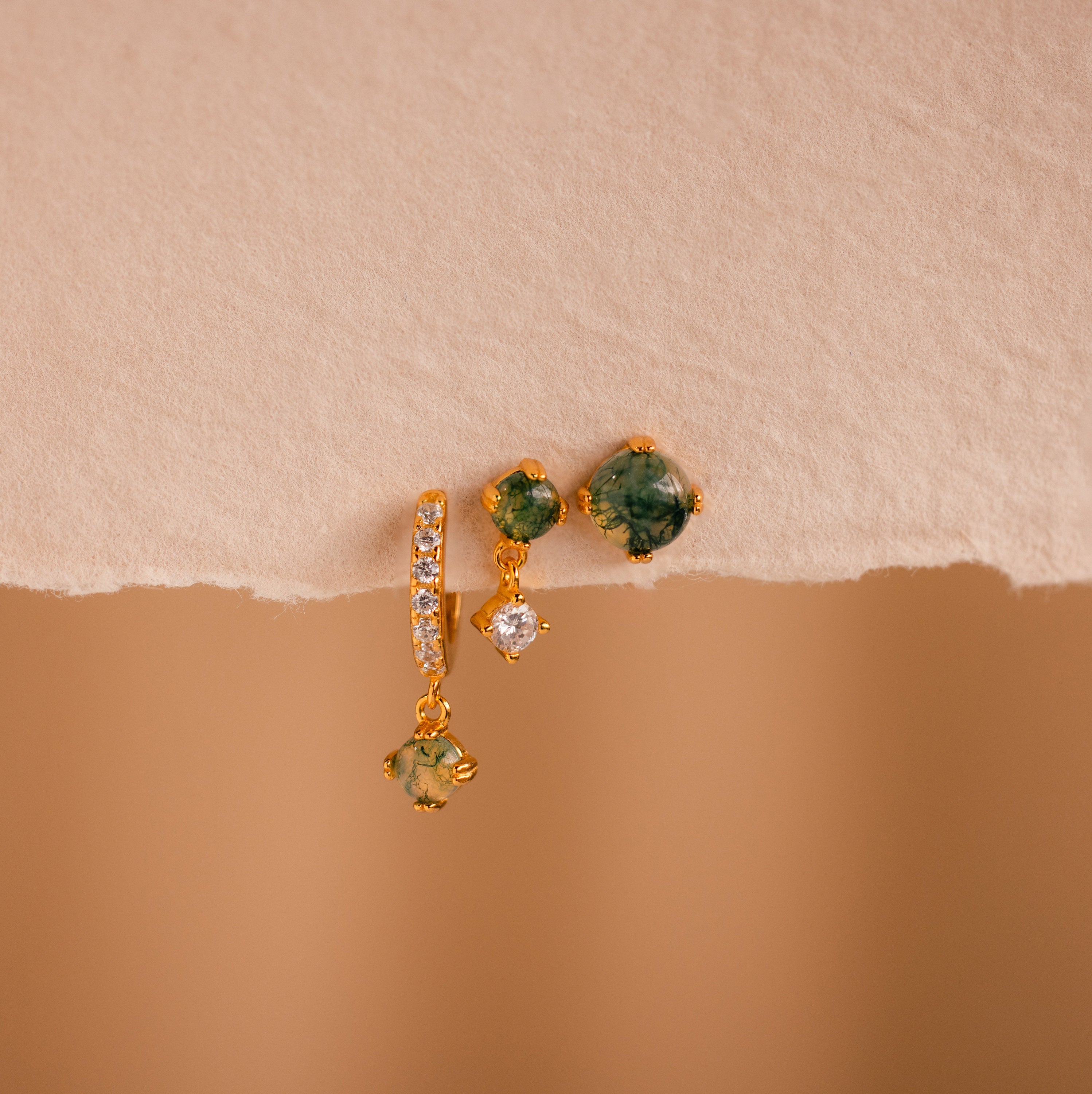 Three gold single earrings from our Agate Earrings Set, adorned with green and clear stones, beautifully displayed on a textured beige surface.