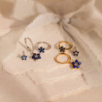 The Sapphire Flower Earrings Set - Silver features gold and silver floral jewelry with blue gemstone accents, beautifully displayed on textured cream paper—an elegant choice for an anniversary gift.