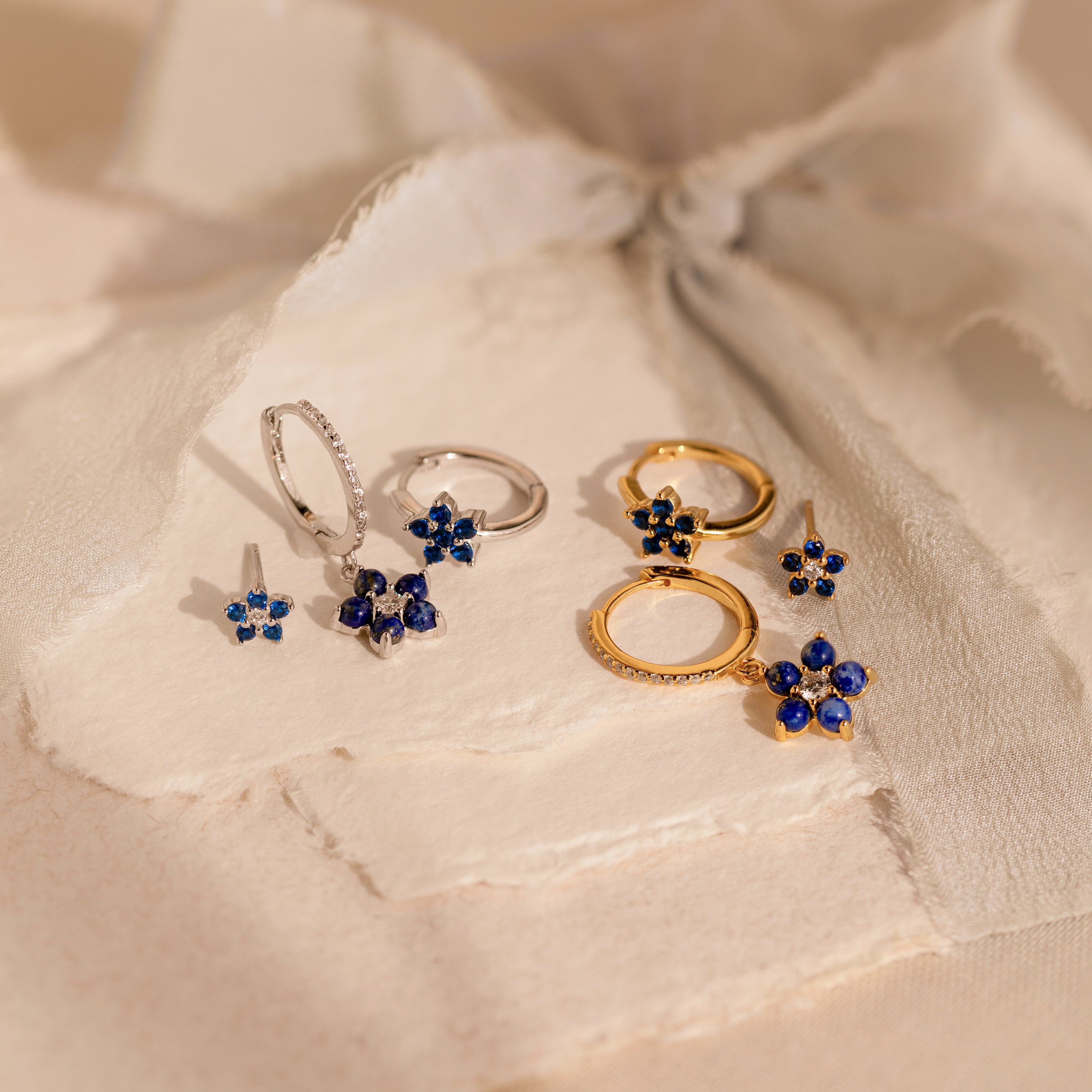 The Sapphire Flower Earrings Set - Silver features gold and silver floral jewelry with blue gemstone accents, beautifully displayed on textured cream paper—an elegant choice for an anniversary gift.