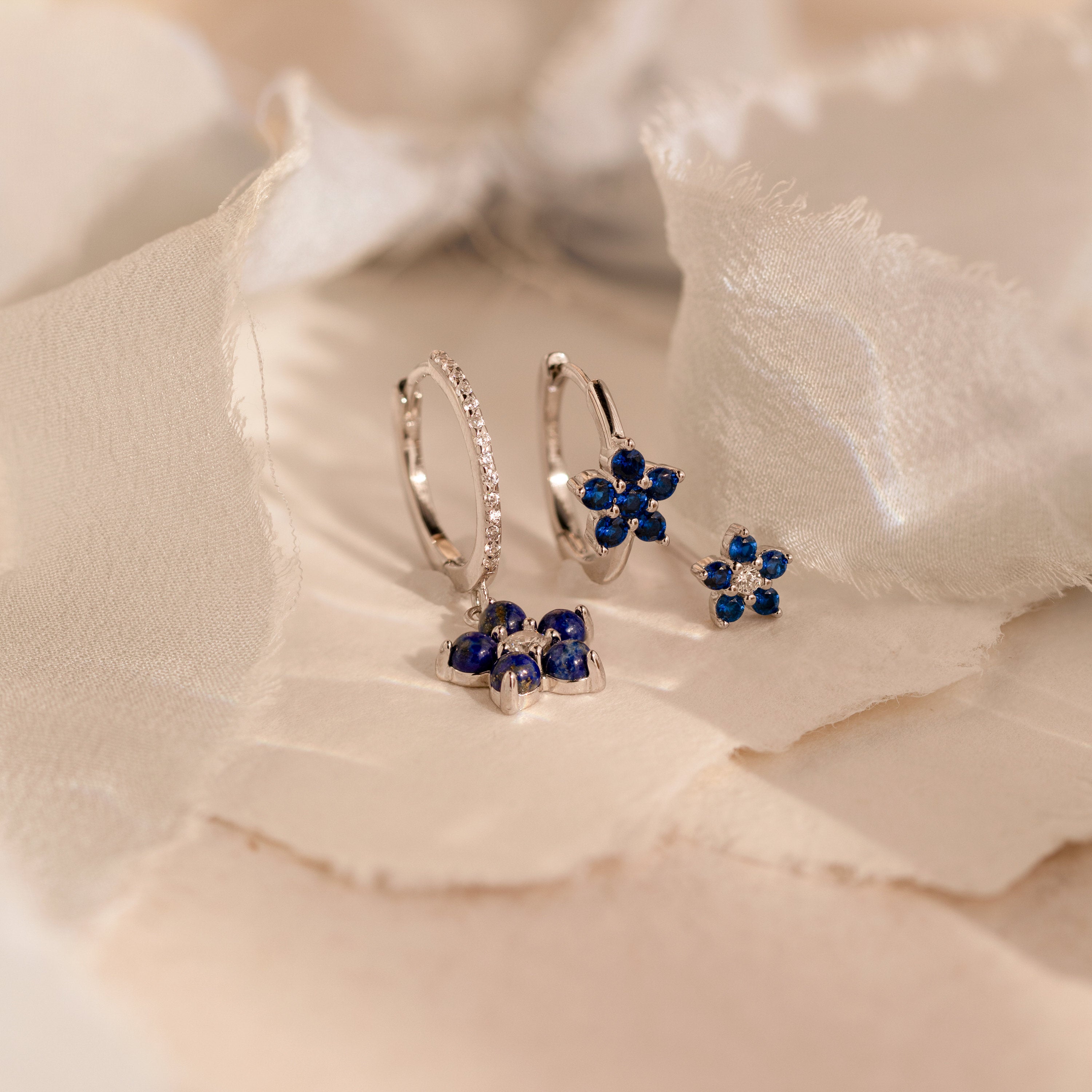 The Sapphire Flower Earrings Set - Silver features two pairs of silver earrings with blue gemstone flower designs on a soft, beige textured background—an elegant anniversary gift.