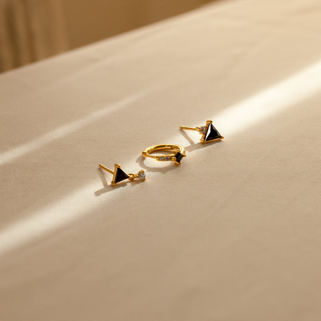 The Black Art Deco Earrings Set, featuring geometric shapes and black stones, rests on beige fabric in sunlight—a striking example of minimalist jewelry.