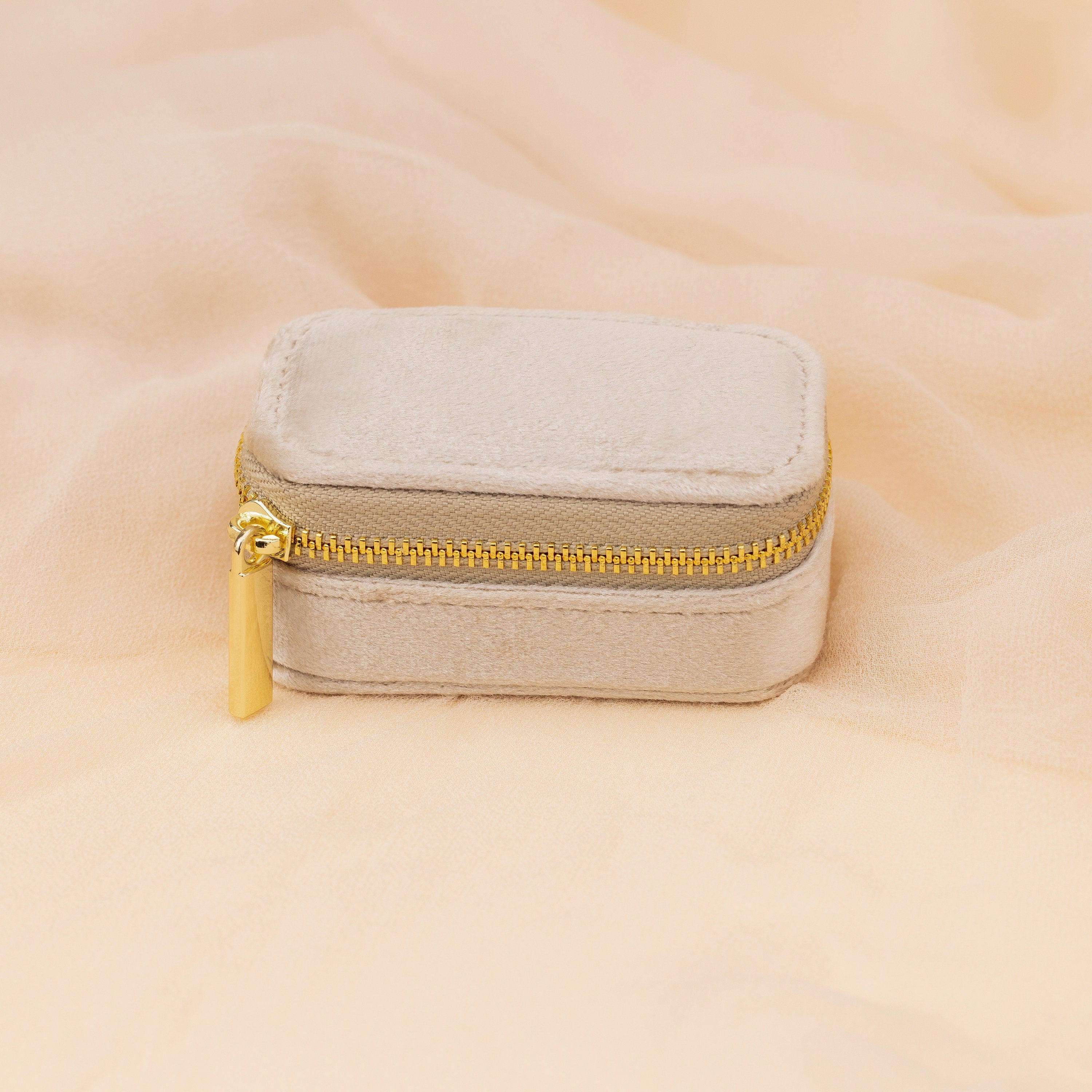 The Mini Travel Jewelry Box in beige velvet, featuring a gold zipper, is displayed on a matching soft fabric background.