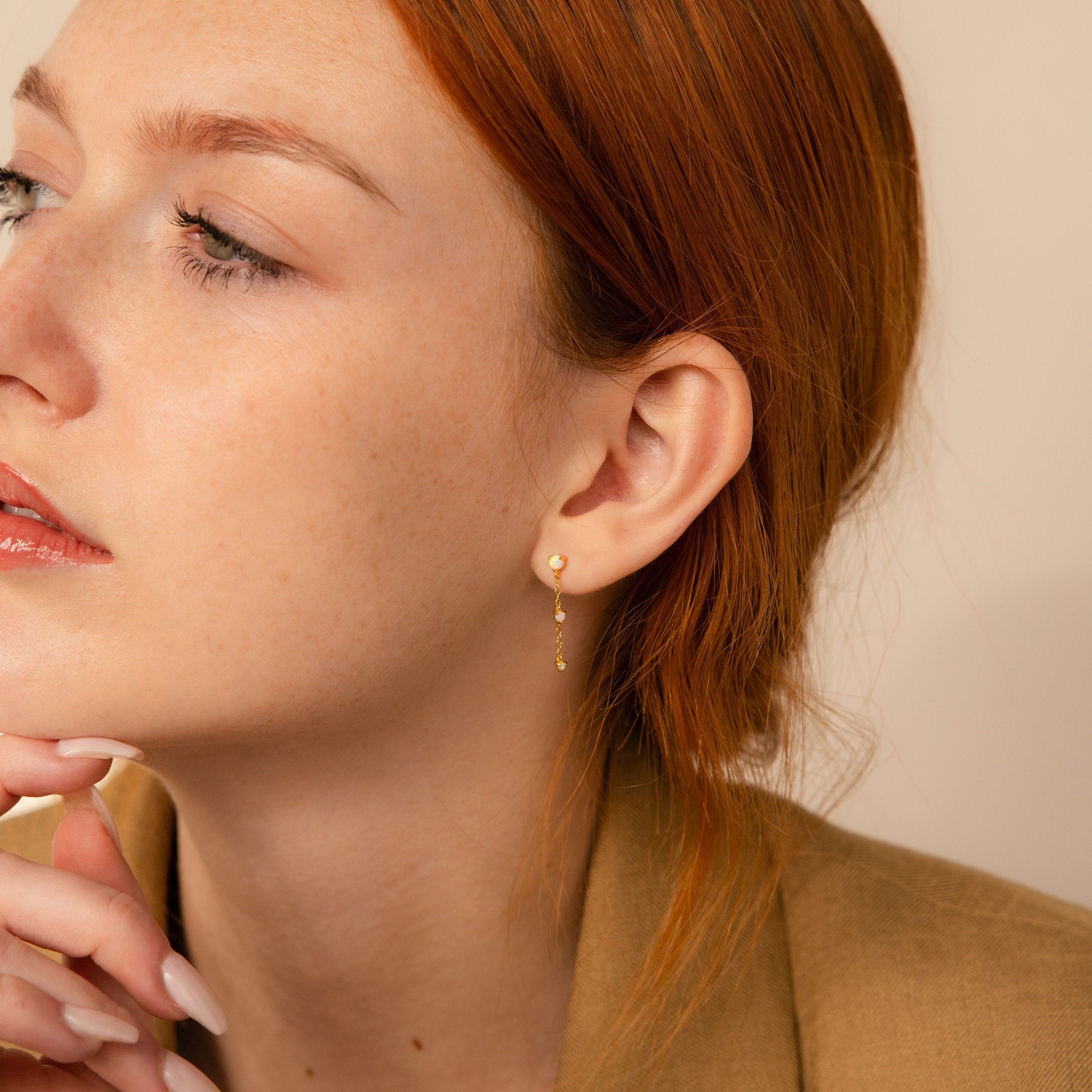 A red-haired woman in a tan blazer gazes thoughtfully to the side, showcasing elegant Opal Chain Drop Earrings.