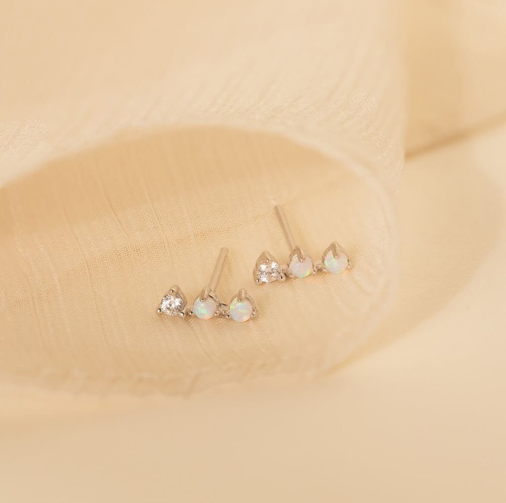 A set of three small stud earrings, featuring Opal Diamond Bar Studs with clear and opal stones, displayed on a soft beige fabric background.