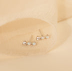A set of three small stud earrings, featuring Opal Diamond Bar Studs with clear and opal stones, displayed on a soft beige fabric background.