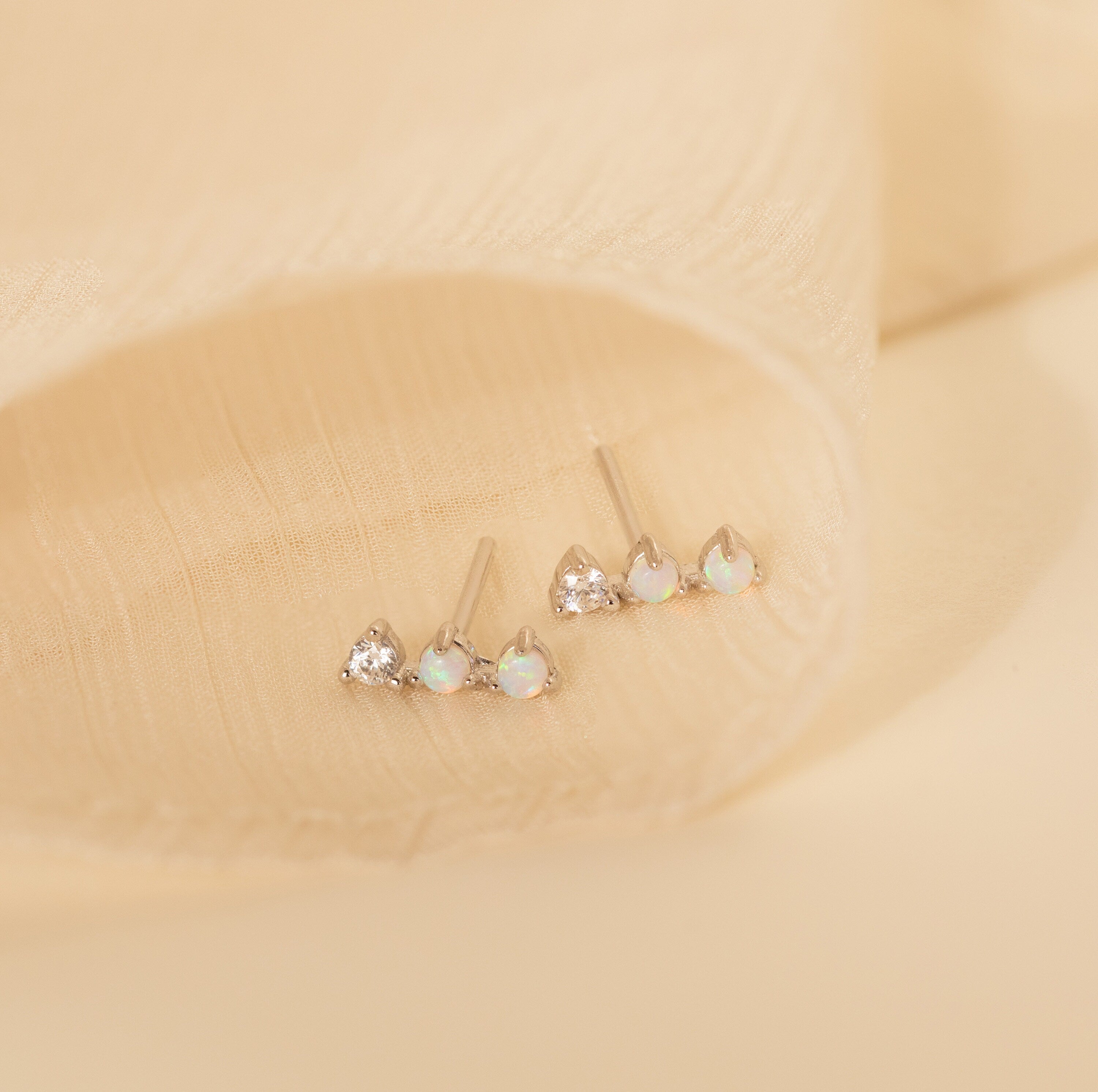 A set of three small stud earrings, featuring Opal Diamond Bar Studs with clear and opal stones, displayed on a soft beige fabric background.