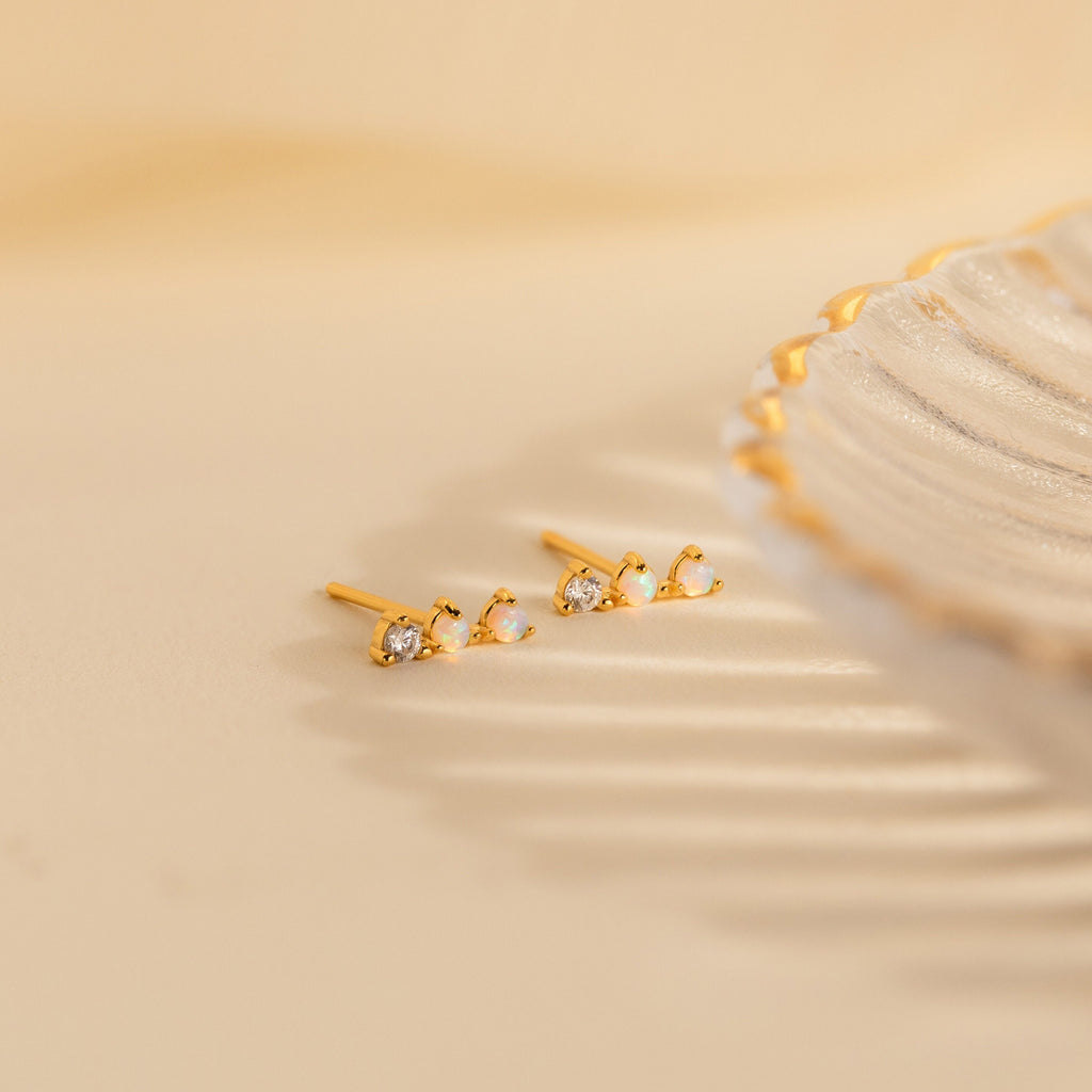 The Opal Diamond Bar Studs, featuring delicate gold settings with sparkling opals and diamonds, rest beside a decorative scalloped-edge dish.