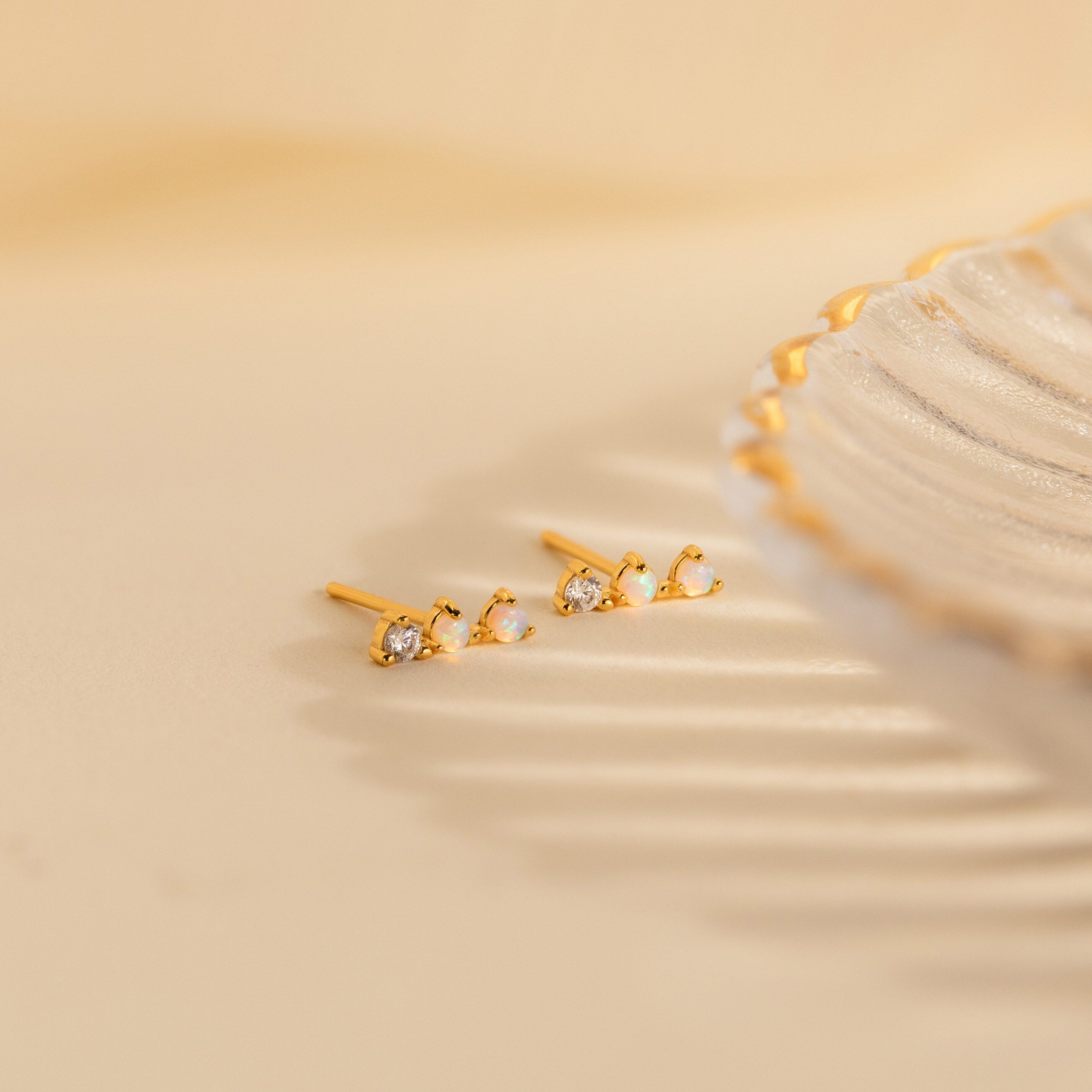 The Opal Diamond Bar Studs, featuring delicate gold settings with sparkling opals and diamonds, rest beside a decorative scalloped-edge dish.